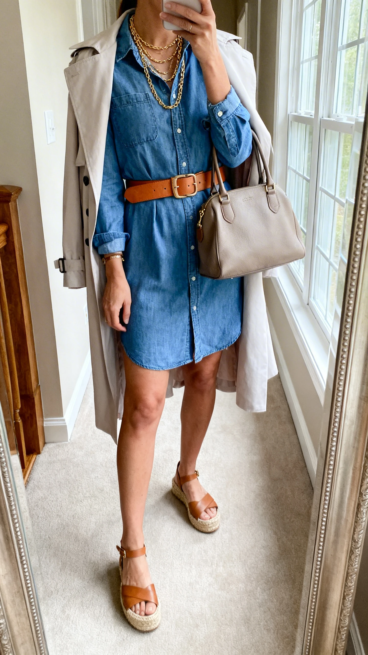 iPhone back-view hallway mirror selfie of a woman wearing a chambray shirt dress cinched with a tan leather belt, espadrille wedges, a light trench coat draped over shoulders, layered gold necklaces, and a medium satchel, face not visible, natural daylight from window, iPhone photo quality.