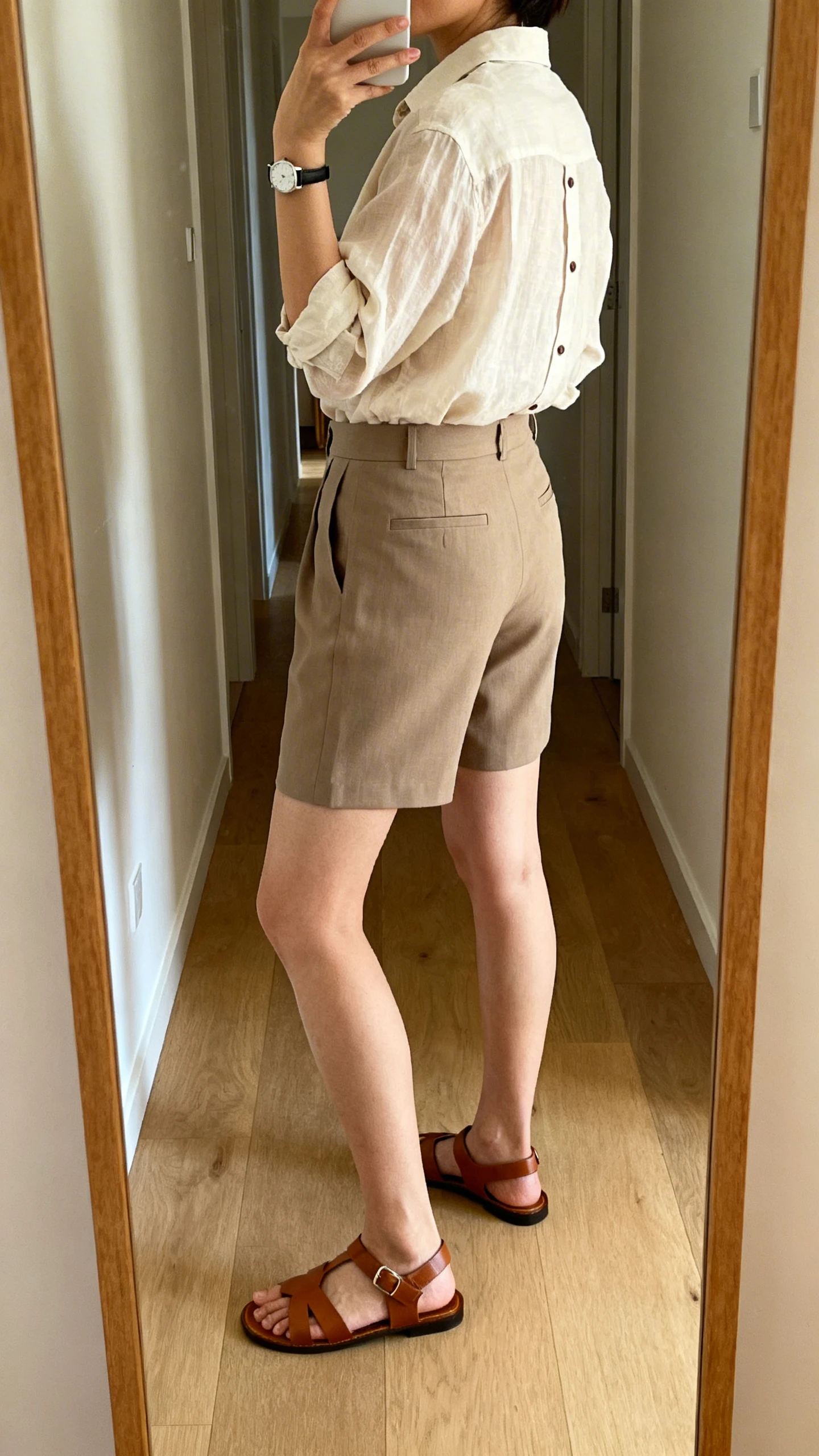 iPhone back-view hallway mirror selfie of a woman wearing an airy button-down tucked into tailored shorts, leather sandals, and a simple watch, face not visible, natural afternoon light, iPhone photo quality.
