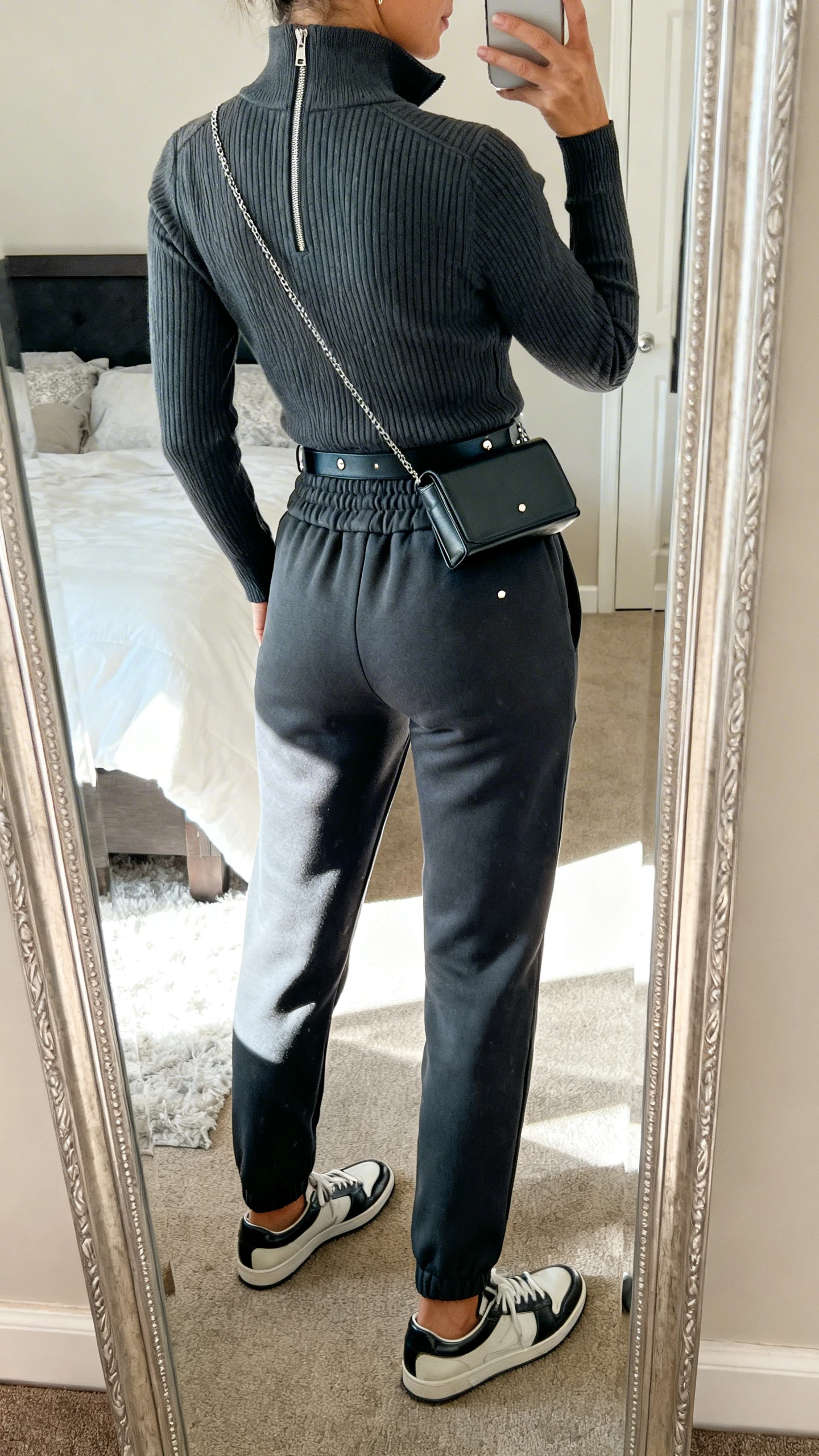iPhone back-view mirror selfie of a woman in a polished quarter-zip knit, tailored joggers with a smooth waistband, clean leather sneakers, a streamlined belt bag, simple studs, and a thin chain, face not visible, bedroom mirror, natural daylight, iPhone photo quality.