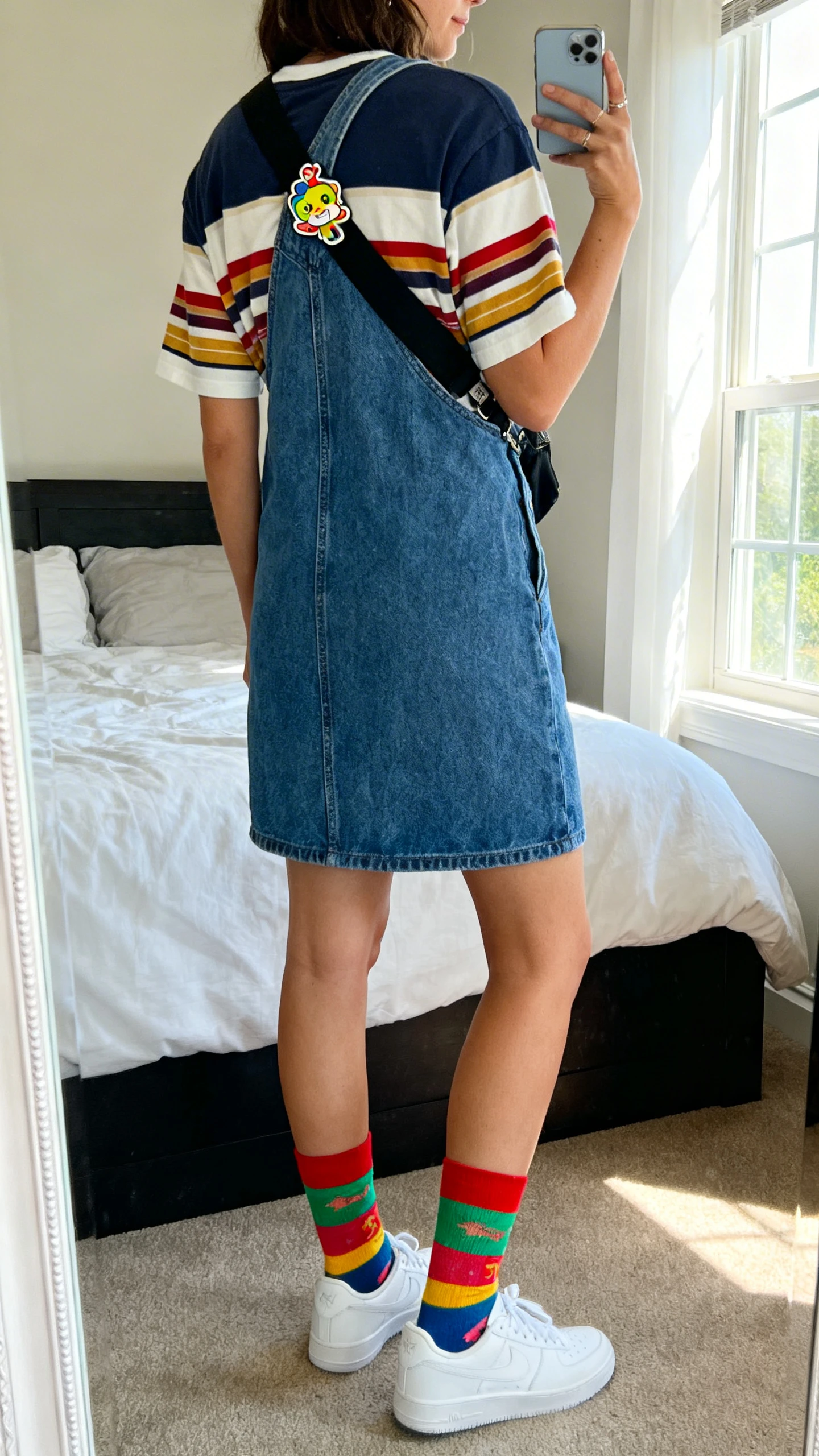 iPhone back view of a woman wearing a layered striped tee under a denim overall dress, colorful crew socks with white sneakers, and a playful enamel pin on the strap, face not visible, casual stance, bedroom mirror, natural window light, iPhone photo quality.