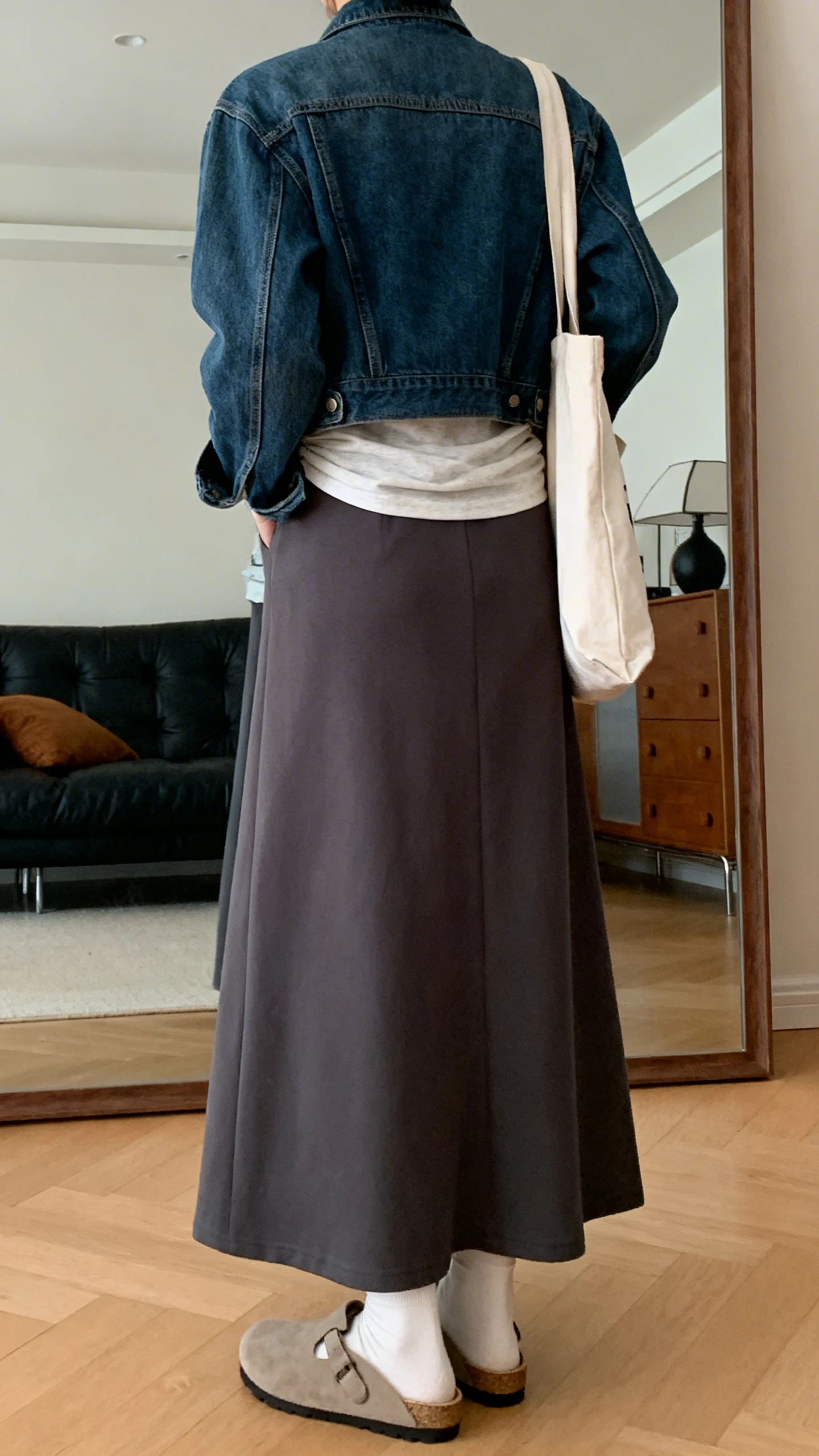 iPhone back view of a woman wearing a maxi skirt with a tucked tee, denim jacket, and comfy clogs, tote bag on shoulder, face not visible, living room mirror, warm indoor lighting, iPhone photo quality.