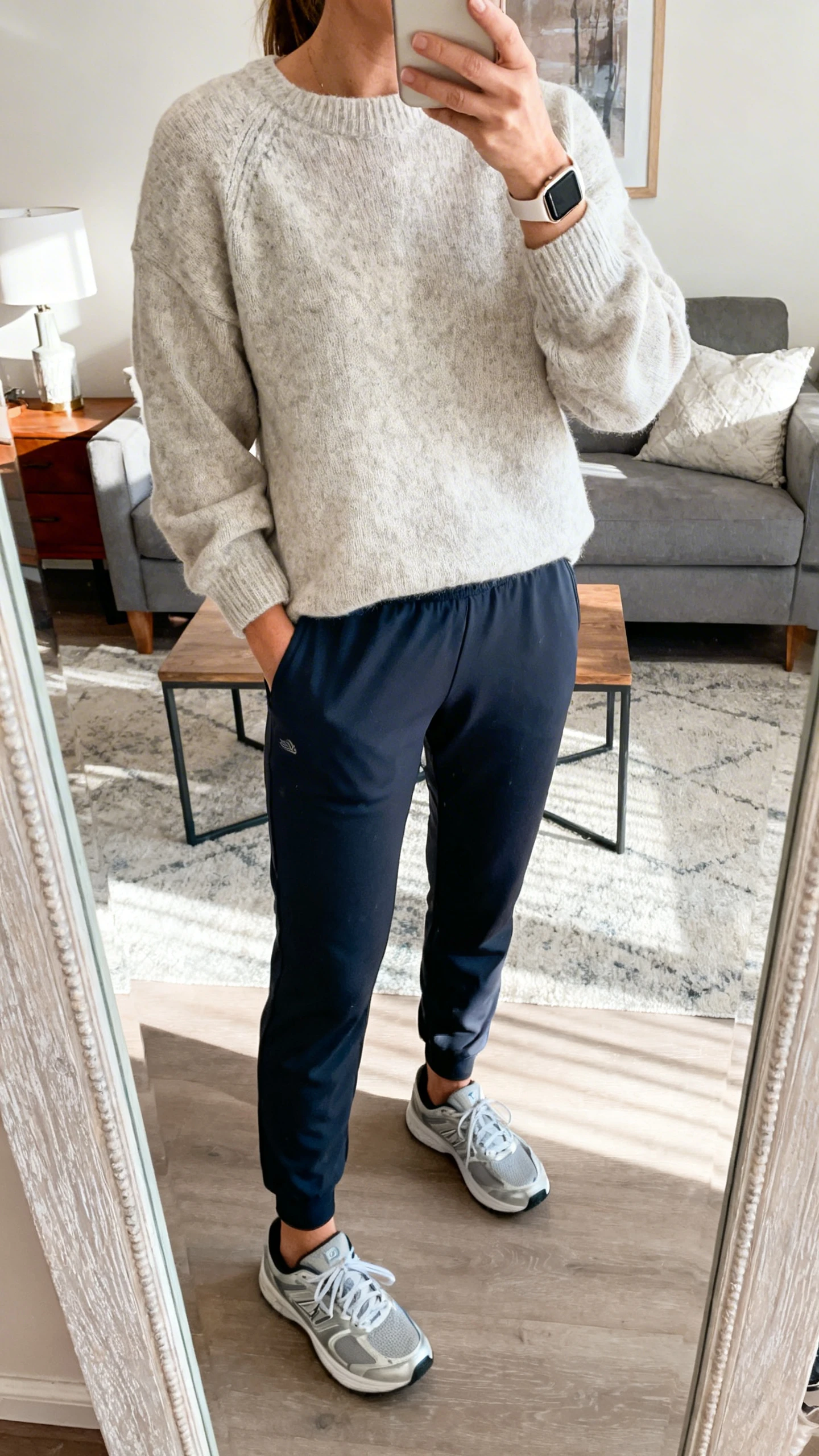 iPhone casual mirror selfie of a woman in a relaxed sweater with ponte joggers and sleek trainers, sporty watch, face not visible, cropped at neck, living-room mirror, morning daylight, iPhone photo quality