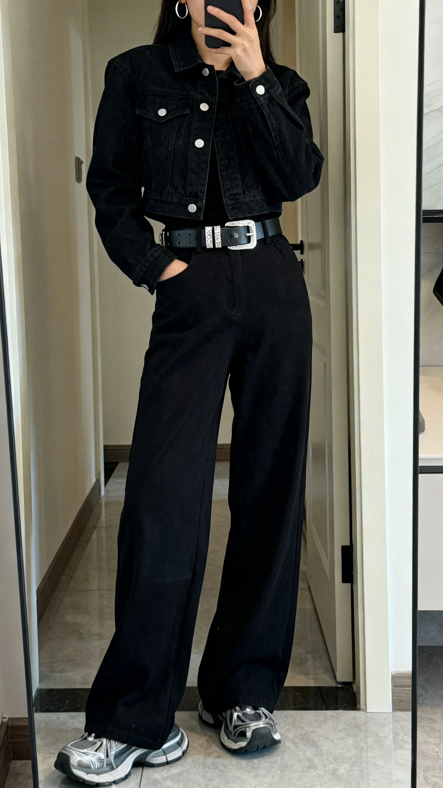 iPhone mirror selfie of a woman in a black denim jacket, wide-leg black pants, bold statement belt with silver hardware, chunky sneakers, minimal hoop earrings, face not visible, cropped at shoulders, full-length mirror in entryway, soft indoor lighting, iPhone photo quality