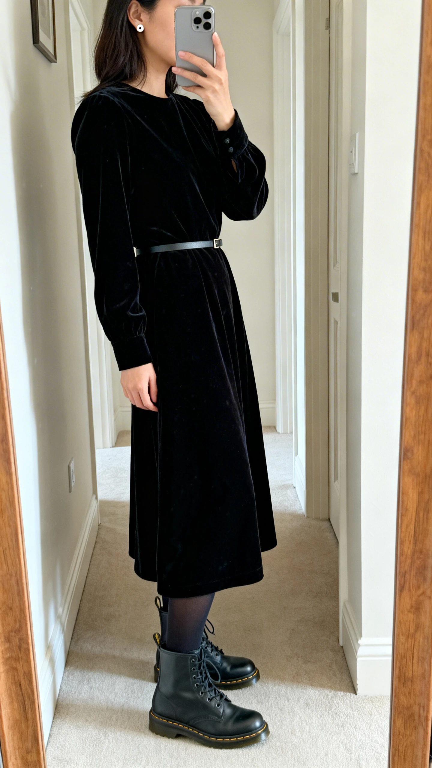 iPhone mirror selfie of a woman in a black velvet midi dress with librarian lace-up boots, dark tights, thin waist belt, simple stud earrings, face not visible, side angle in a hallway mirror, soft indoor lighting, iPhone photo quality