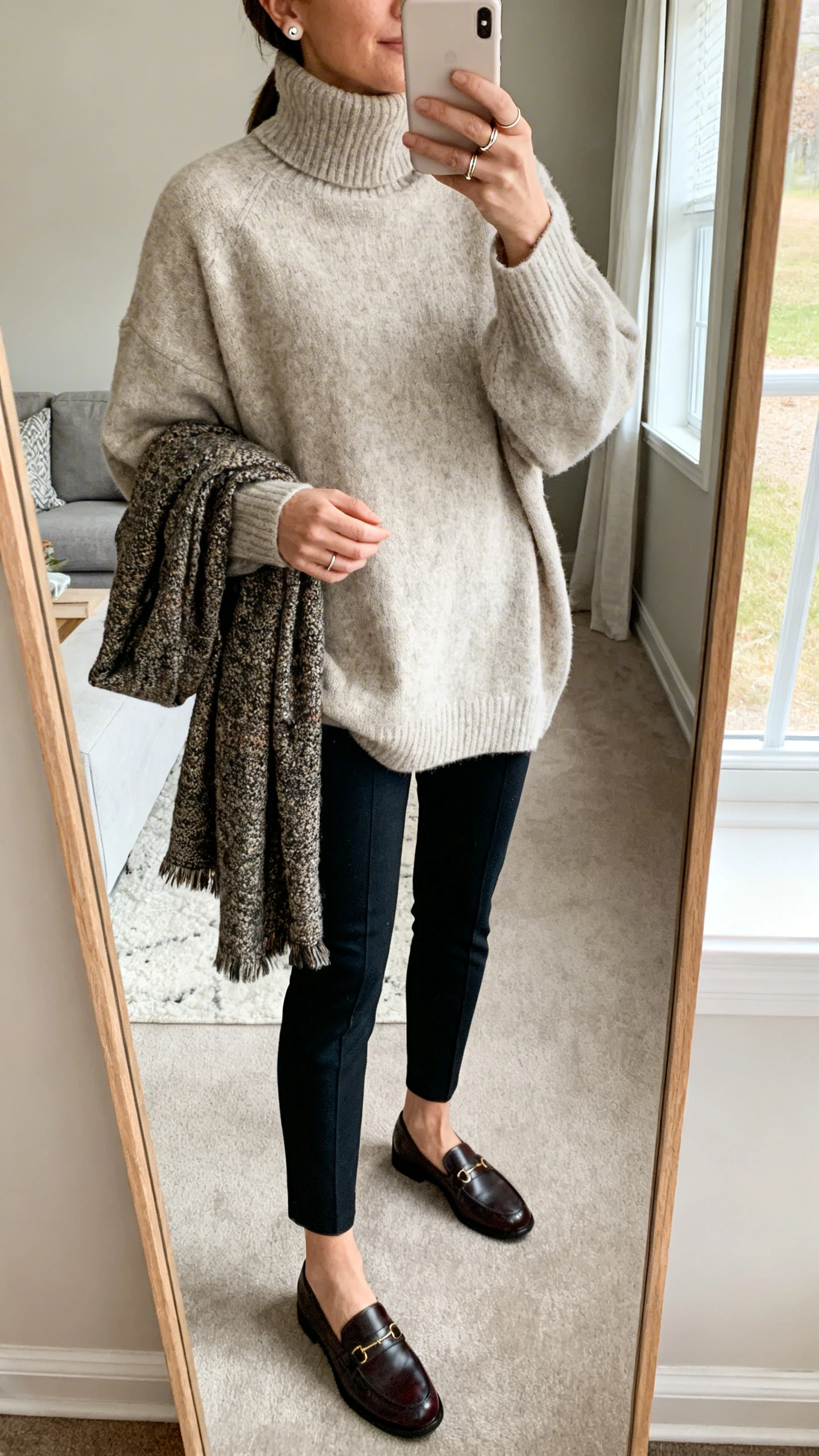 iPhone mirror selfie of a woman in a cozy oversized turtleneck sweater, slim ankle pants, classic loafers, a textured scarf draped loosely, stud earrings, and a dainty ring stack, face not visible, cropped at shoulders, living room mirror, morning window light, iPhone photo quality.