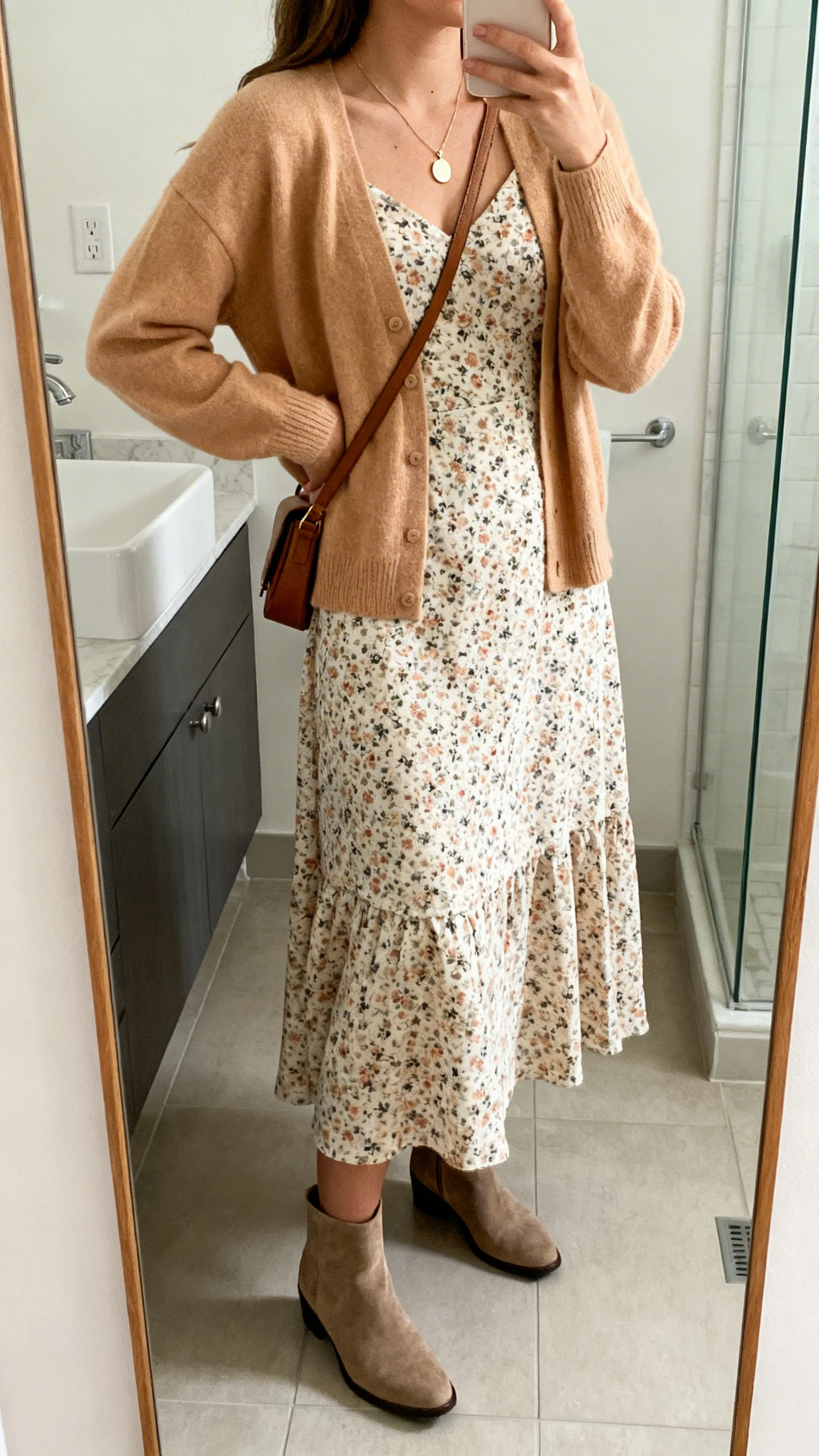 iPhone mirror selfie of a woman in a floral midi dress with a soft tan cardigan layered on top, delicate pendant necklace, ankle boots, and a crossbody bag, face not visible, side angle, bathroom full-length mirror, natural indoor light, iPhone photo quality.
