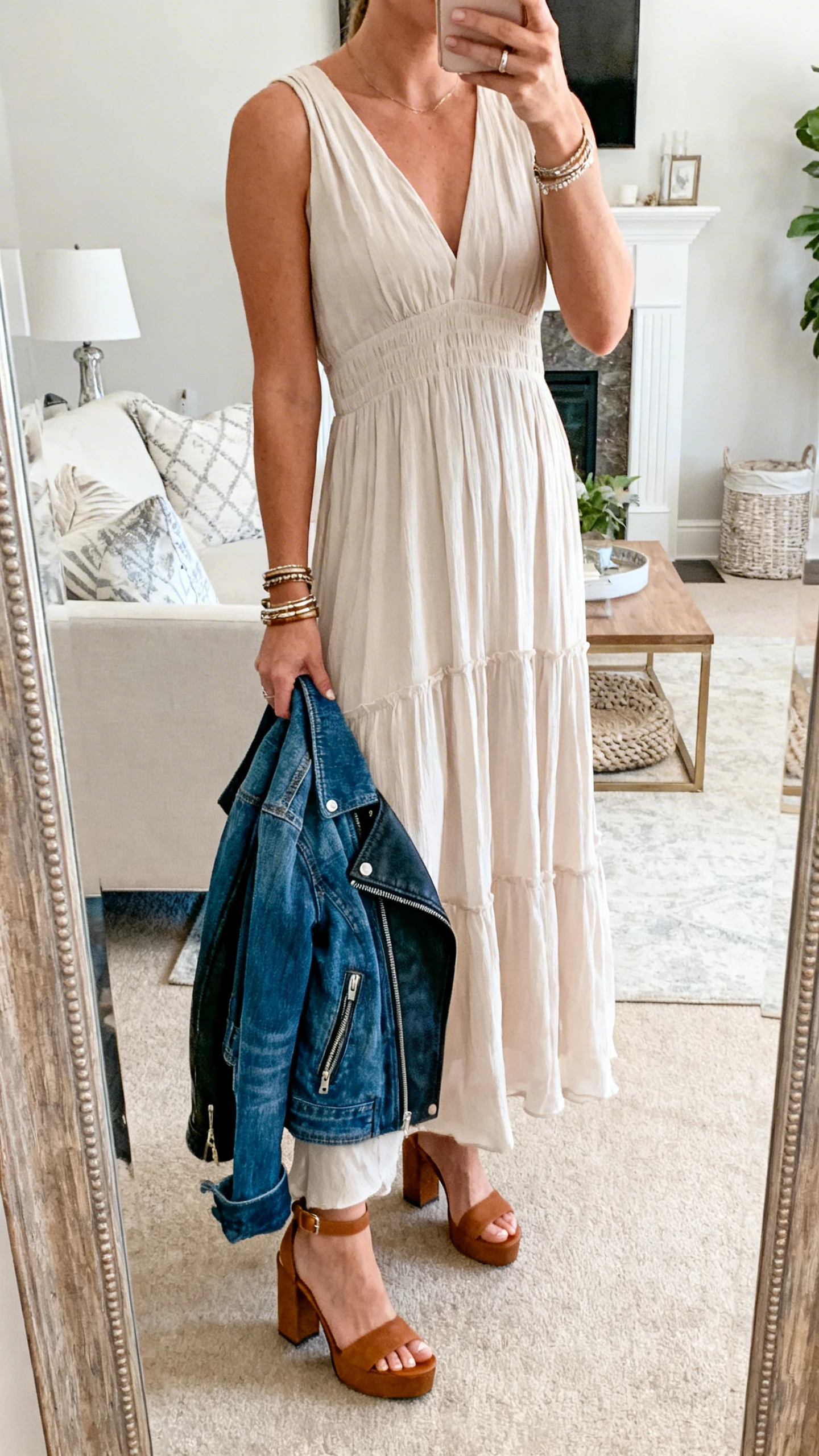 iPhone mirror selfie of a woman in a flowy maxi dress with a denim jacket (optional moto jacket draped over arm), block-heel sandals, and layered bracelets, face not visible, cropped at neck, living room mirror, soft natural daylight, iPhone photo quality.