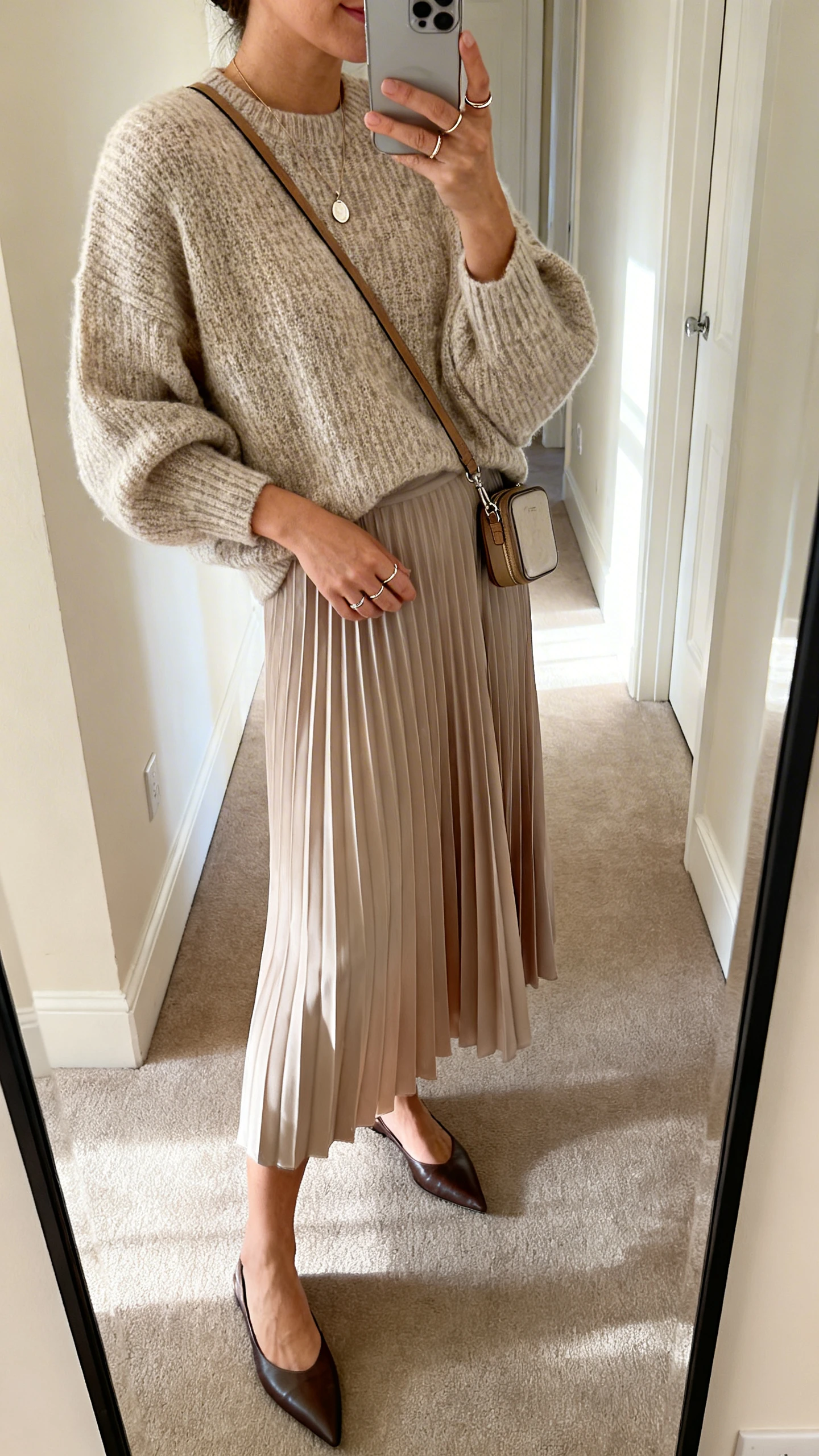 iPhone mirror selfie of a woman in a flowy pleated midi skirt with a boxy lightweight knit sweater, pointed flats, delicate pendant necklace, thin stacking rings, and a compact crossbody, face not visible, side angle in a hallway full-length mirror, morning light, iPhone photo quality.