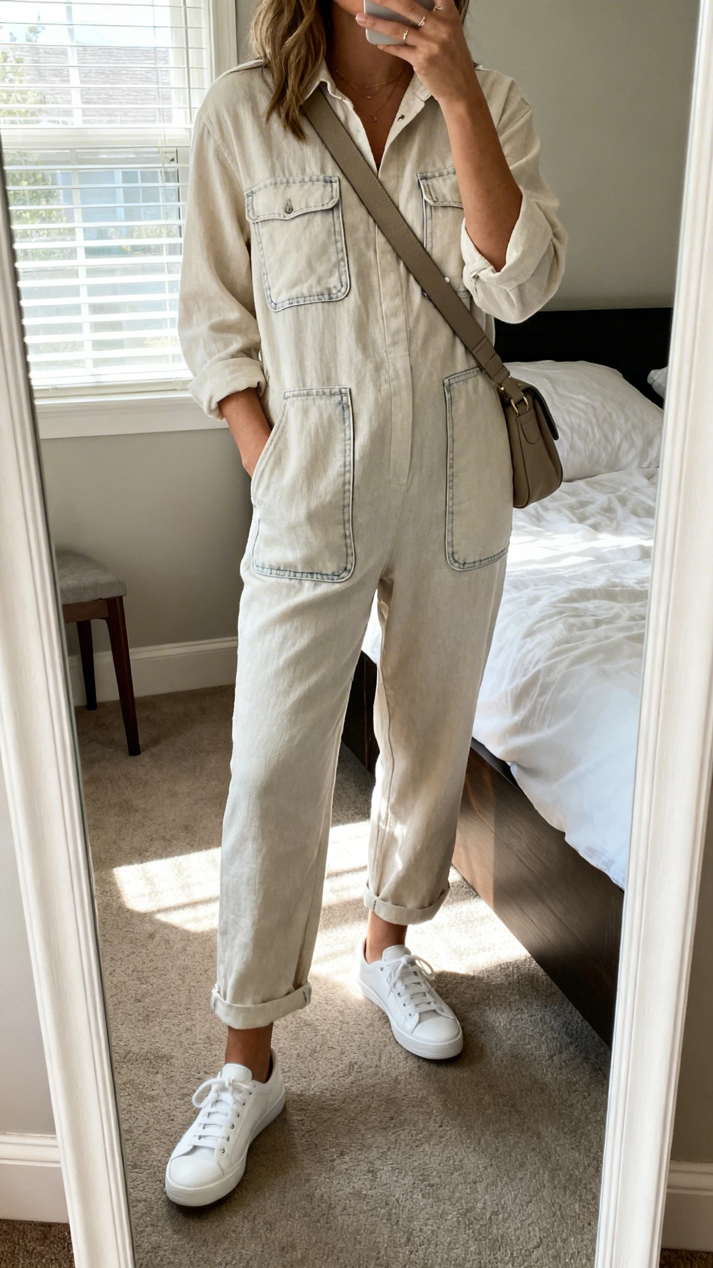 iPhone mirror selfie of a woman in a lightweight jumpsuit with pockets, white low-top sneakers, and a crossbody bag, face not visible, cropped at shoulders, bedroom mirror, morning window light, iPhone photo quality.