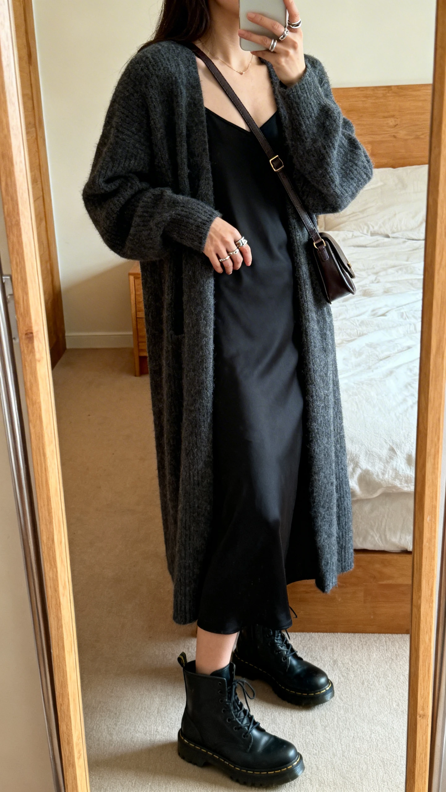 iPhone mirror selfie of a woman in a longline charcoal cardigan over a black midi slip dress, black combat boots, layered rings, crossbody satchel, face not visible, side angle, bedroom mirror, warm indoor lighting, iPhone photo quality