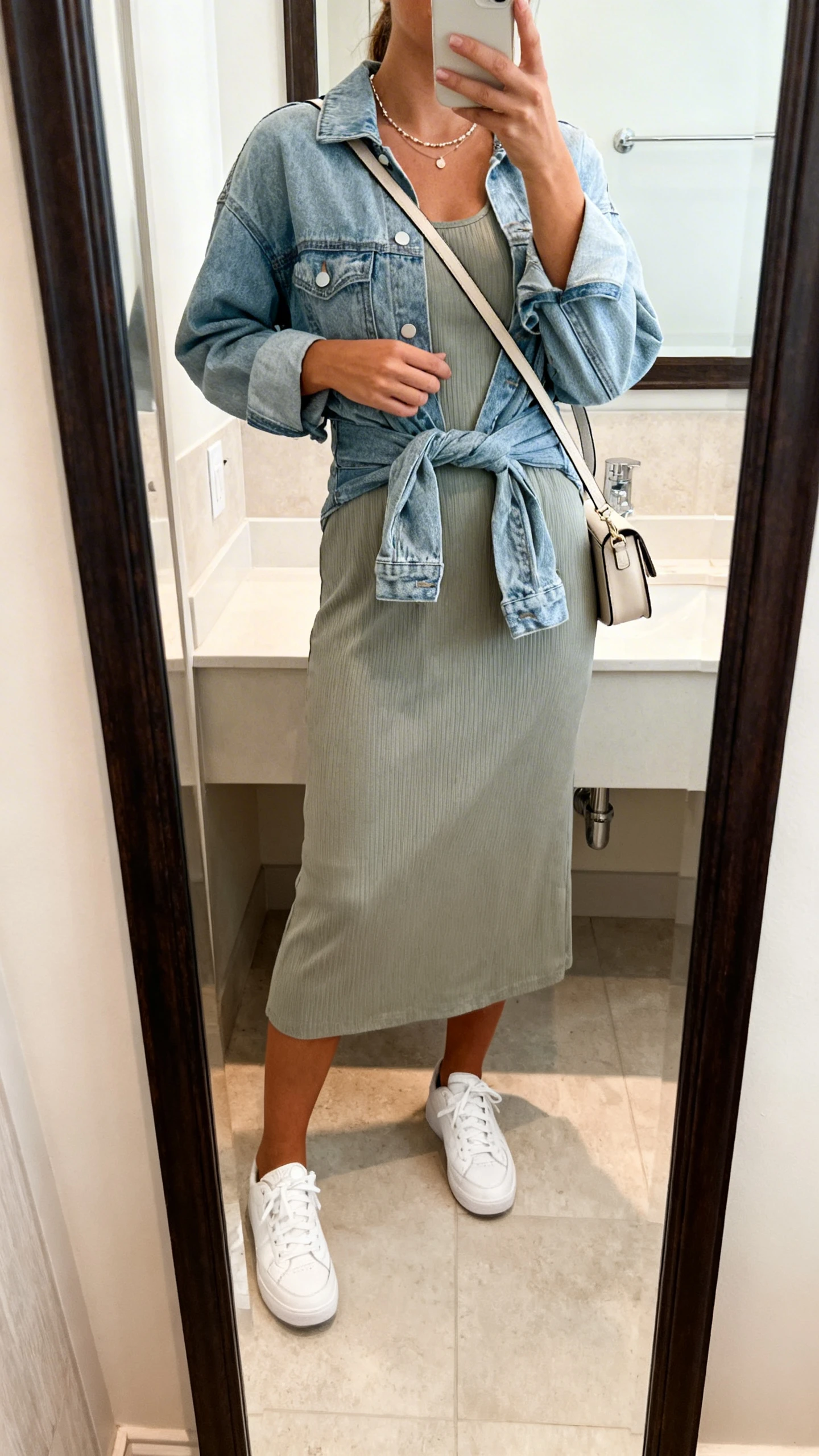 iPhone mirror selfie of a woman in a midi dress with clean white sneakers, crossbody bag, delicate necklace, and lightweight denim jacket tied at the waist, face not visible, bathroom full-length mirror, soft indoor lighting, iPhone photo quality.