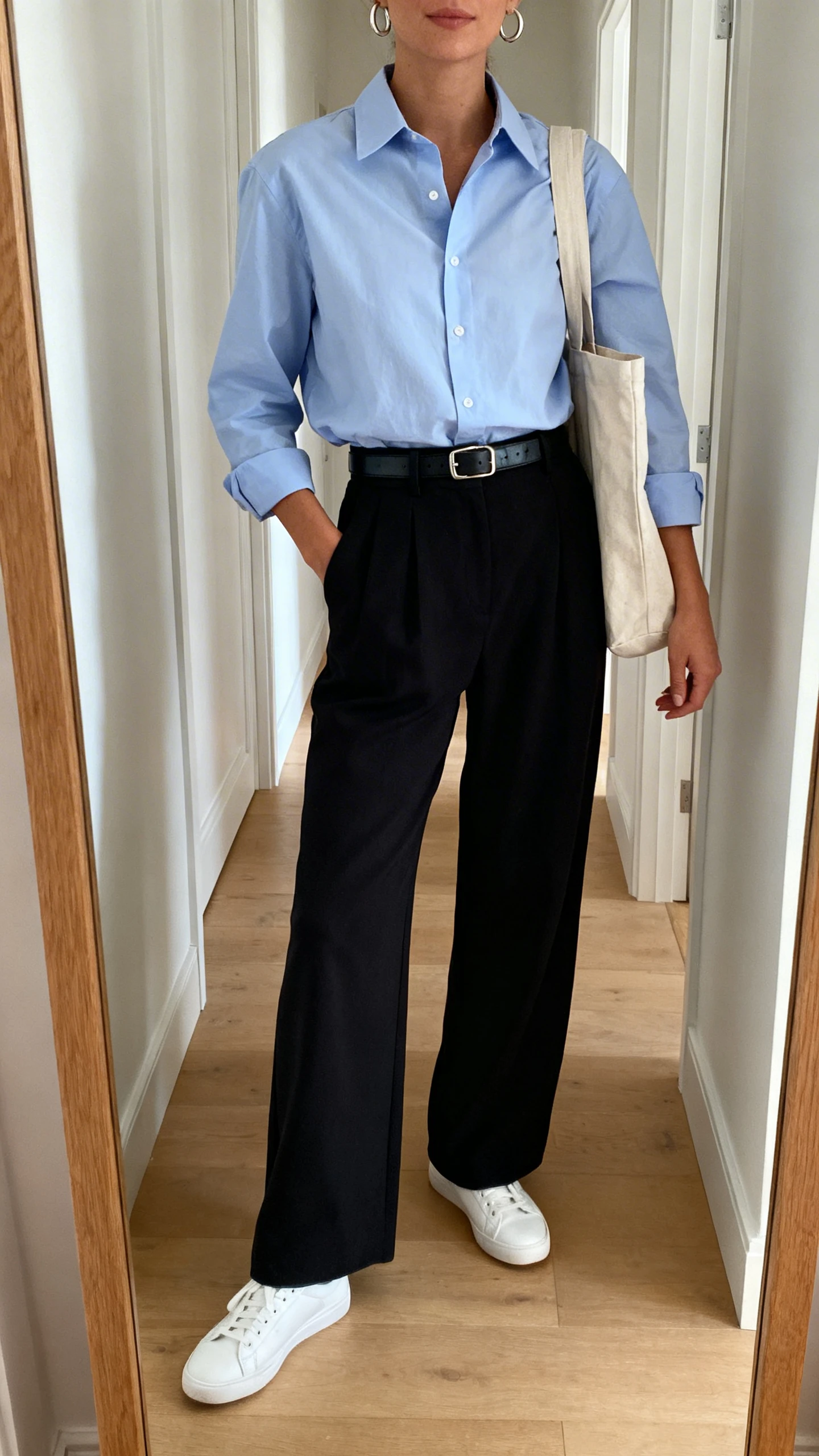 iPhone mirror selfie of a woman in a pale blue cotton oxford shirt tucked into high-rise black wide-leg pants, polished white sneakers, slim leather belt, simple hoop earrings, and a canvas tote, face not visible, cropped at shoulders, full-length hallway mirror, natural daylight, iPhone photo quality.