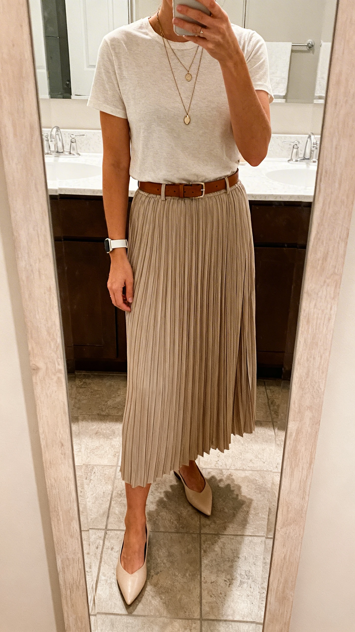 iPhone mirror selfie of a woman in a pleated midi skirt (solid or micro-print) with a slightly structured high-quality cotton/modal t-shirt, pointed-toe flats, a slim watch, layered pendant necklace, and an optional leather belt, face not visible, bathroom mirror, natural indoor lighting, iPhone photo quality.