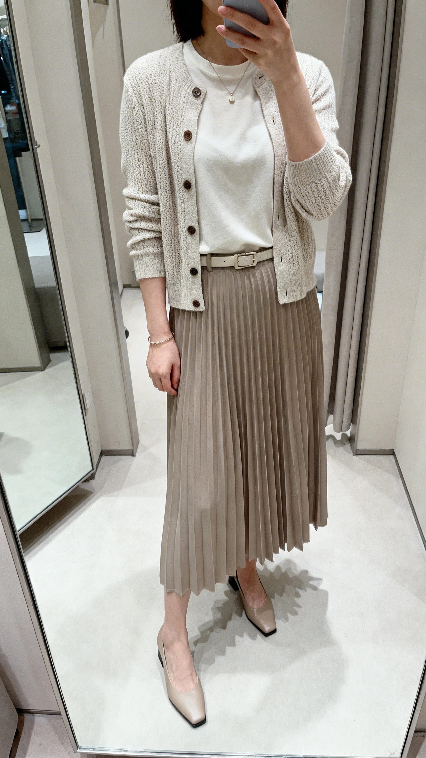 iPhone mirror selfie of a woman in a pleated midi skirt, fine-knit cardigan buttoned as a top over a simple crewneck tee, low block heels, a subtle belt, a small pendant, and a delicate bracelet, face not visible, cropped at neck, dressing-room style mirror, soft morning light, iPhone photo quality.