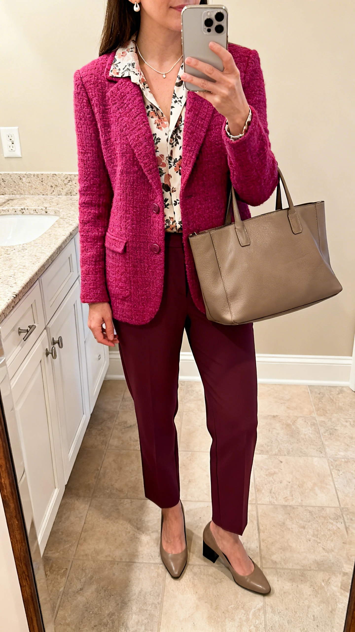 iPhone mirror selfie of a woman in a saturated-color knit blazer, a printed blouse, tailored cigarette pants, low block-heel pumps, a fine necklace with understated earrings, and a roomy tote, face not visible, cropped at shoulders, bathroom mirror, warm indoor lighting, iPhone photo quality.