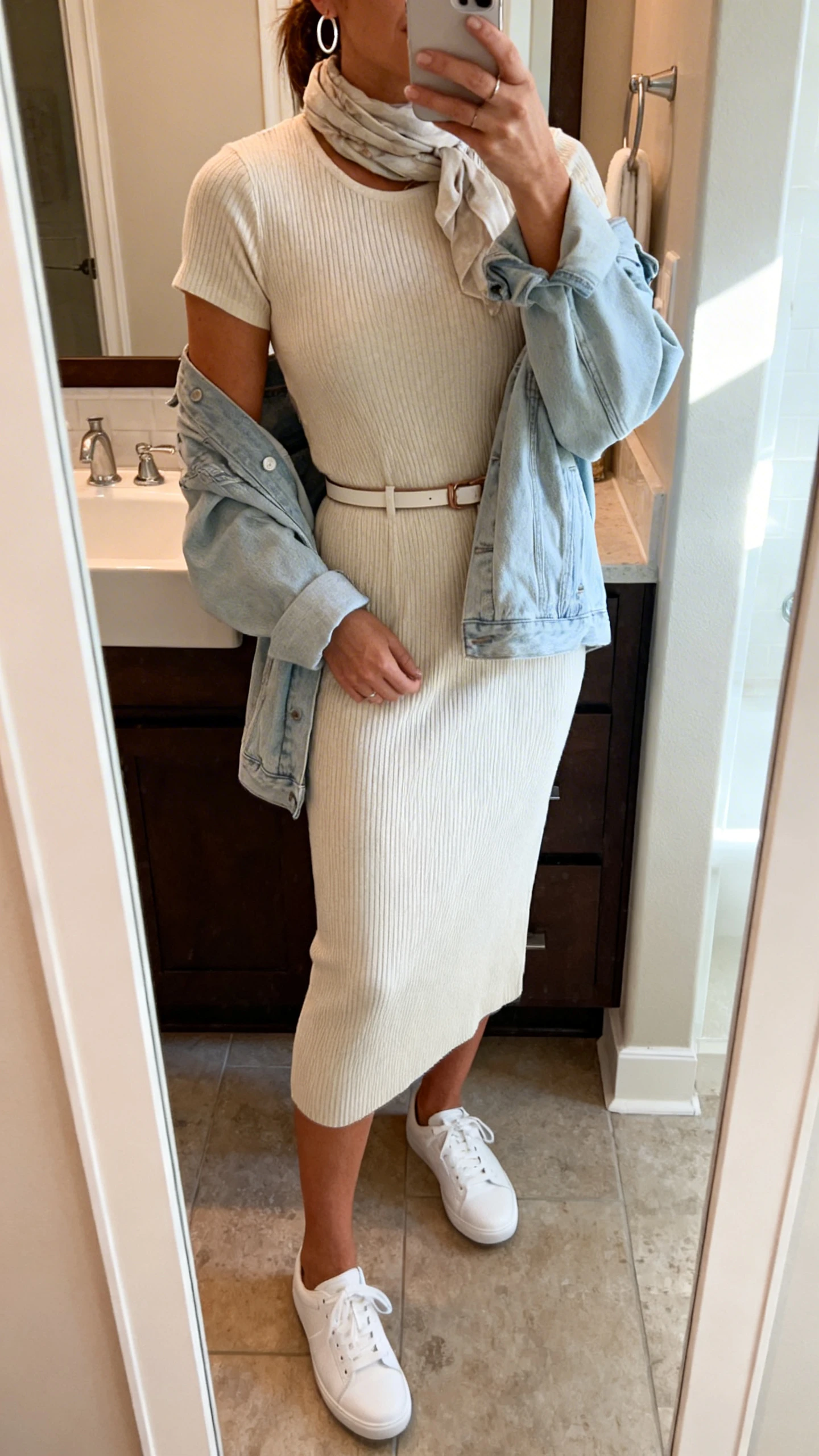 iPhone mirror selfie of a woman in a soft ribbed knit midi dress (short sleeve) with clean white low-profile sneakers, a light denim jacket draped over shoulders, simple hoop earrings, thin belt, and a lightweight scarf, face not visible, bathroom mirror, indoor natural light, iPhone photo quality.