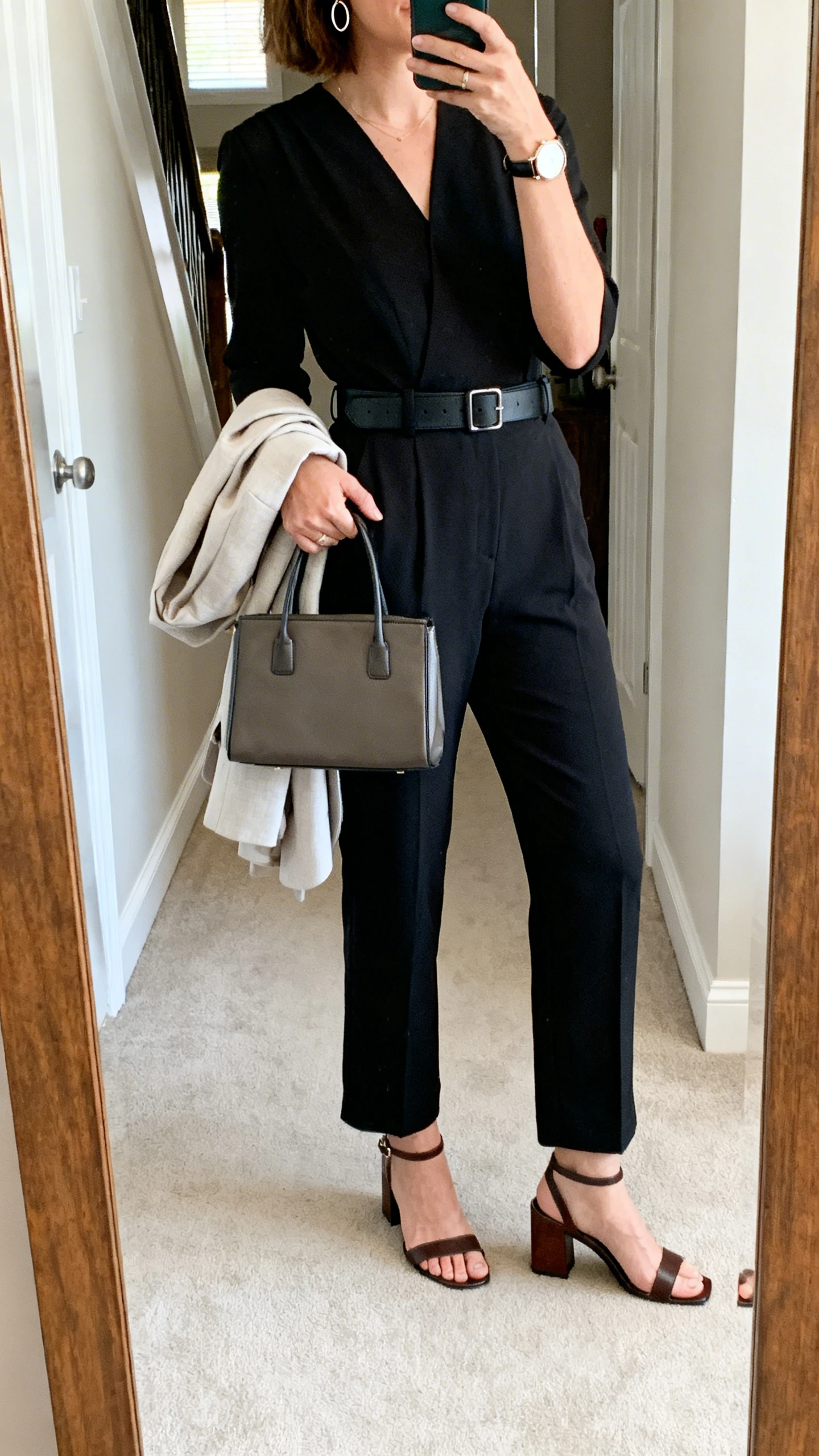 iPhone mirror selfie of a woman in a tailored black jumpsuit with a comfortable waist-defining belt, block-heel sandals, minimal hoops, a watch, a structured tote, and a light blazer over her arm, face not visible, cropped at shoulders, entryway full-length mirror, indoor daylight, iPhone photo quality.