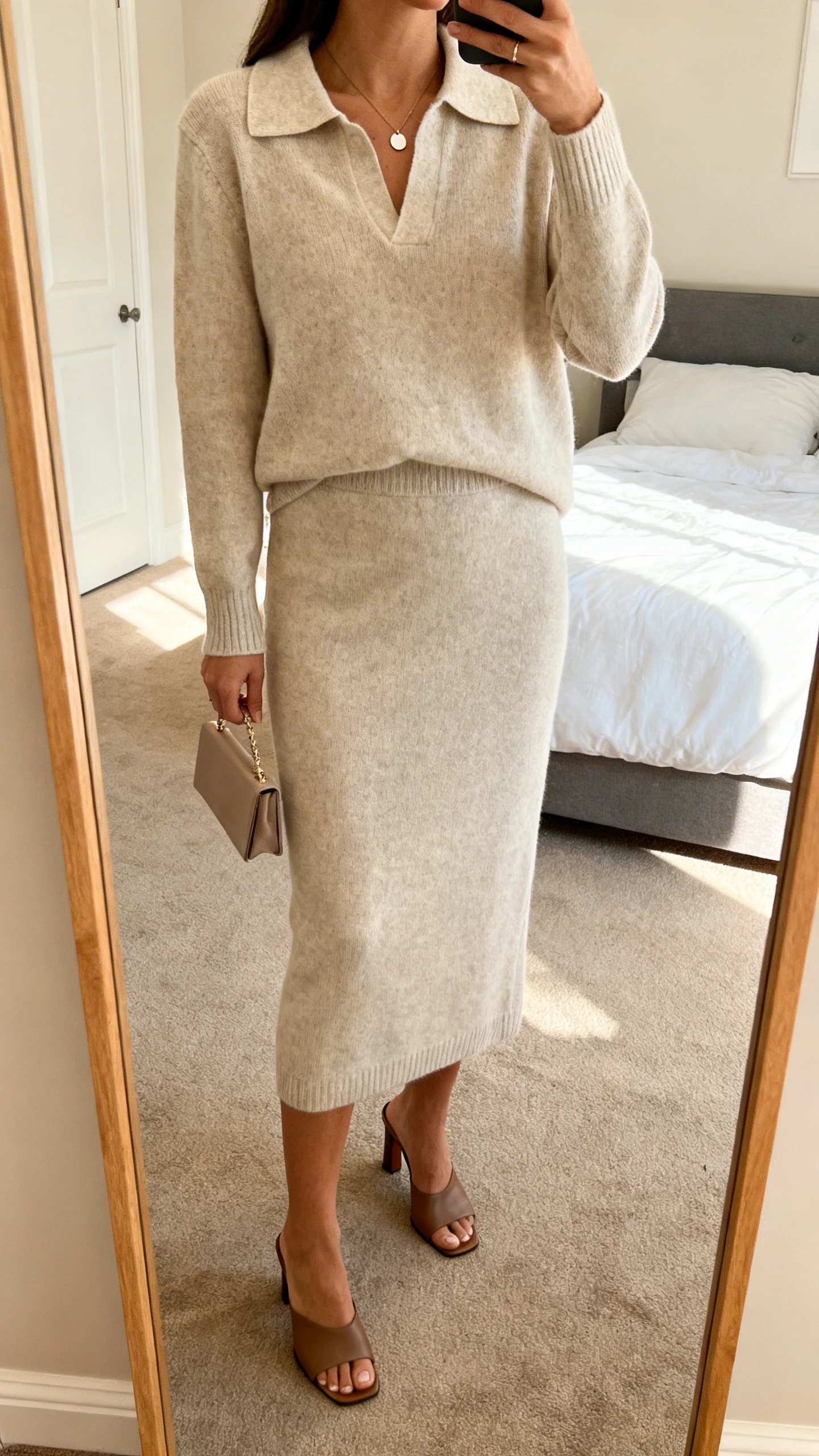iPhone mirror selfie of a woman in an elevated knit set (collared knit top and matching midi skirt) with heeled mules, minimal pendant, and petite clutch, face not visible, cropped at shoulders, bedroom full-length mirror, warm indoor lighting, iPhone photo quality.