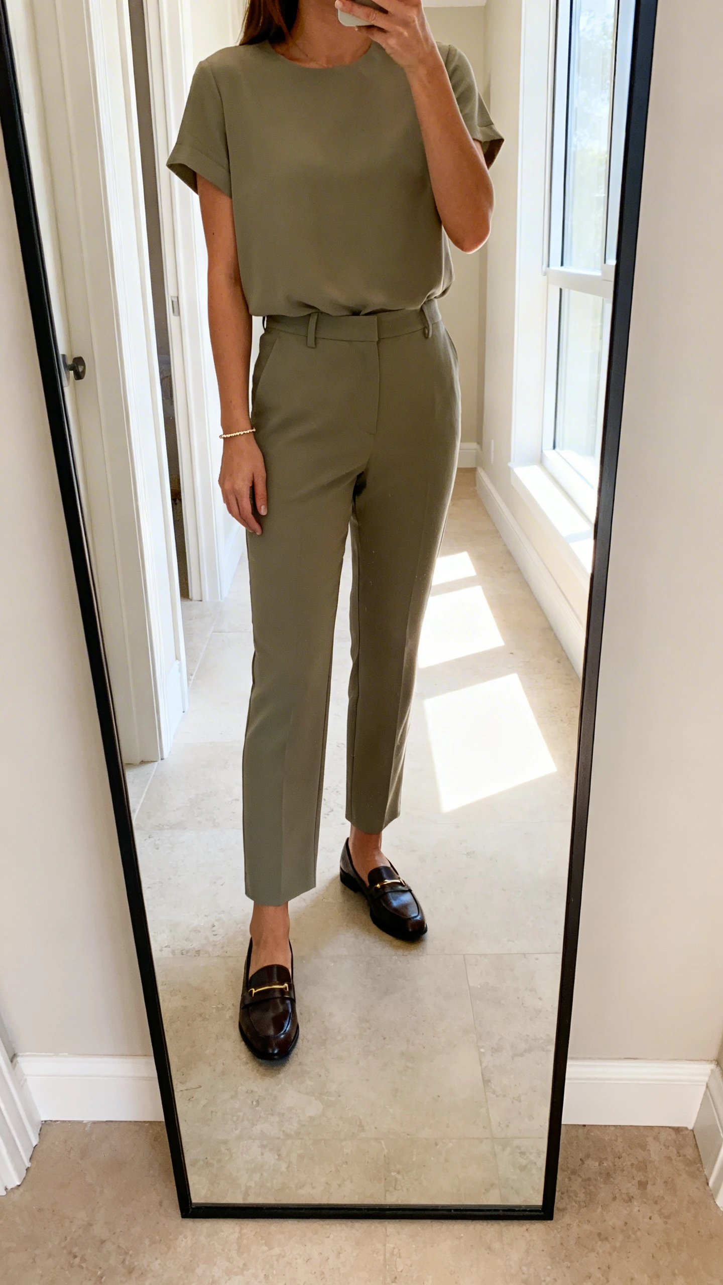 iPhone mirror selfie of a woman in an elevated matching set (short-sleeve top and coordinated trousers) with sleek loafers and a delicate bracelet, face not visible, cropped at shoulders, full-length mirror in hallway, bright window light, iPhone photo quality.