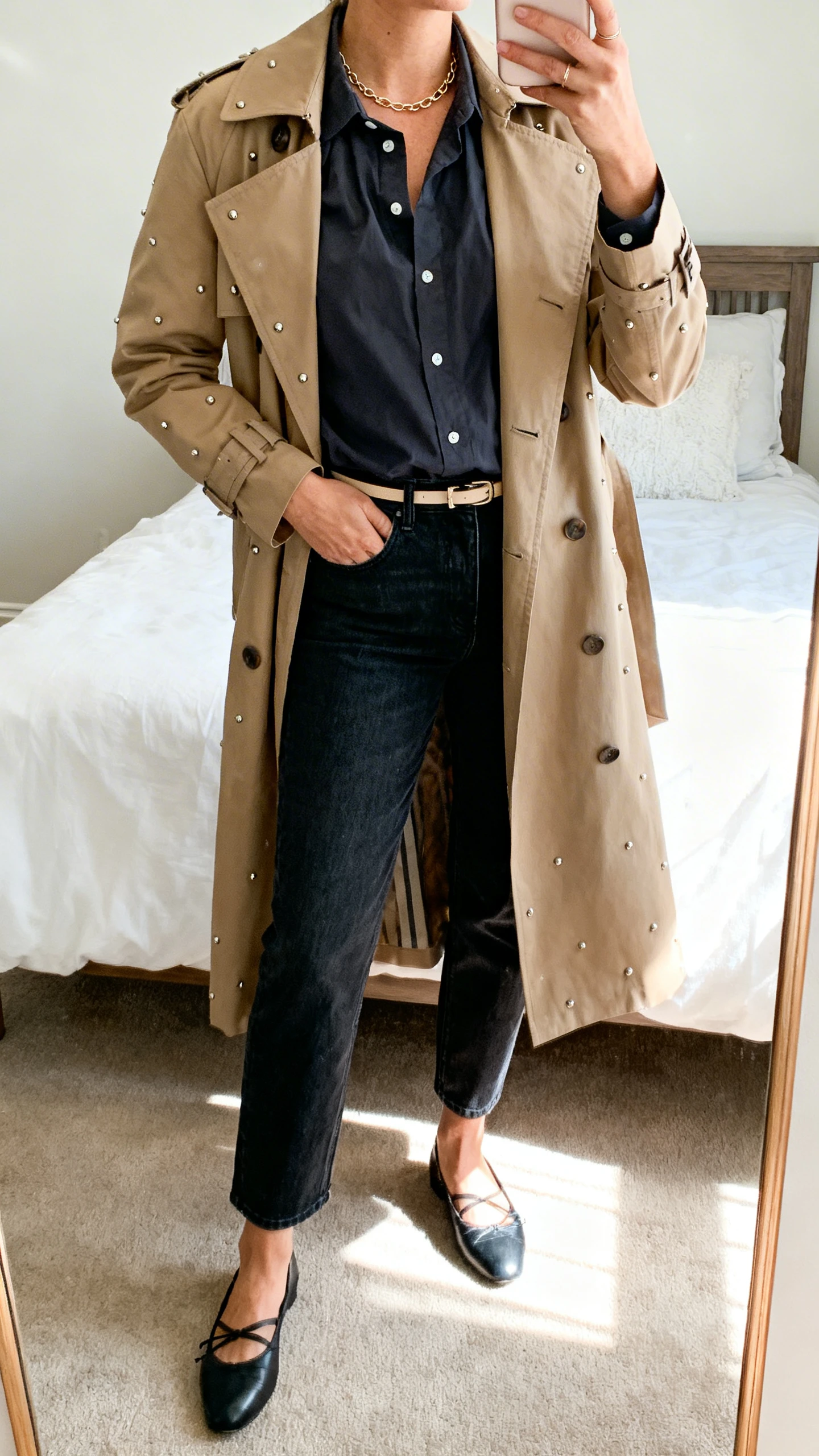 iPhone mirror selfie of a woman in dark-wash straight-leg jeans, a structured button-up poplin shirt, polished ballet flats, a thin belt, and a classic trench coat with subtle studs and a dainty chain necklace, face not visible, bedroom mirror, bright natural light, iPhone photo quality.
