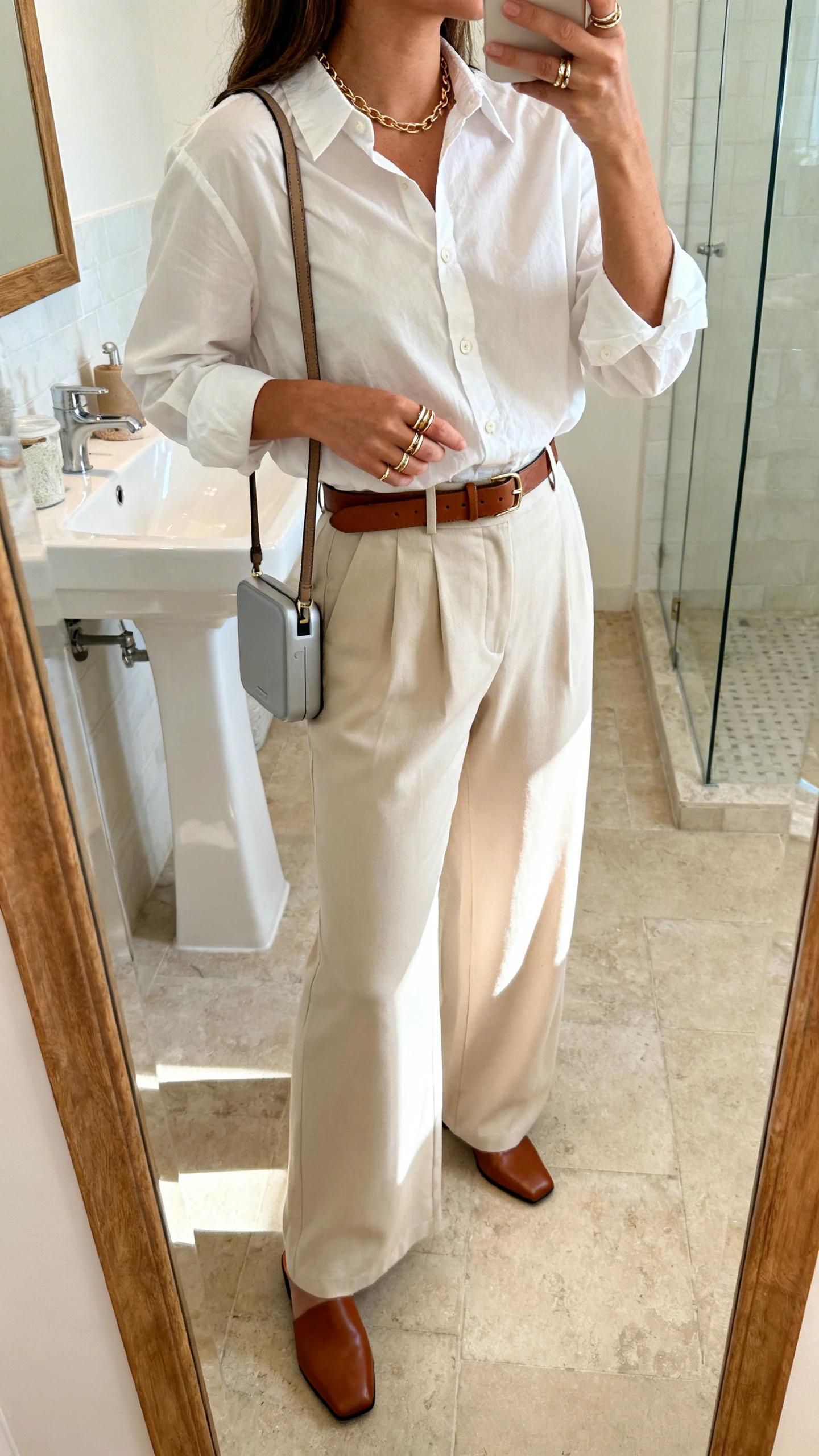 iPhone mirror selfie of a woman in high-rise wide-leg trousers (lightweight) with a crisp poplin button-up, polished mules, delicate chain necklace, stackable rings, optional leather belt, and a portable crossbody, face not visible, side angle in a bathroom mirror with bright indoor light, iPhone photo quality.