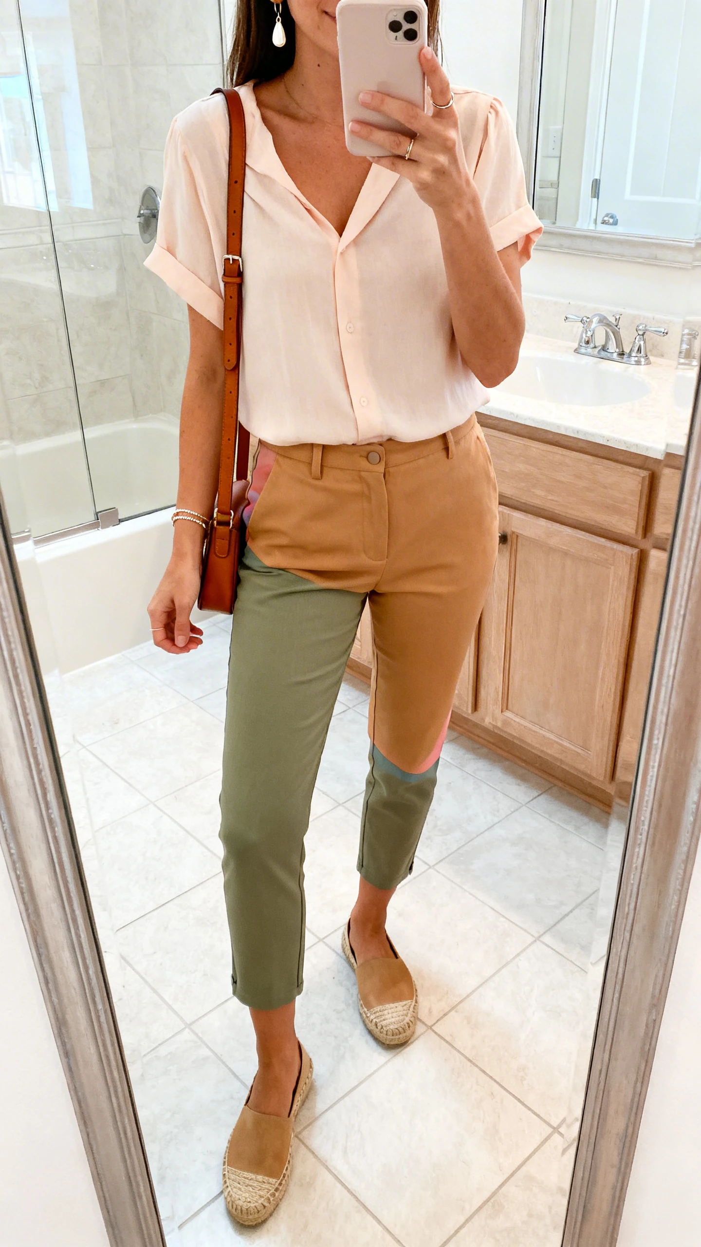 iPhone mirror selfie of a woman in slim cropped chinos (tan/olive/pastel) with a soft short-sleeve blouse, comfortable espadrilles, simple drop earrings, a delicate bracelet, and a leather crossbody, face not visible, cropped at neck in a bathroom mirror with bright indoor light, iPhone photo quality.