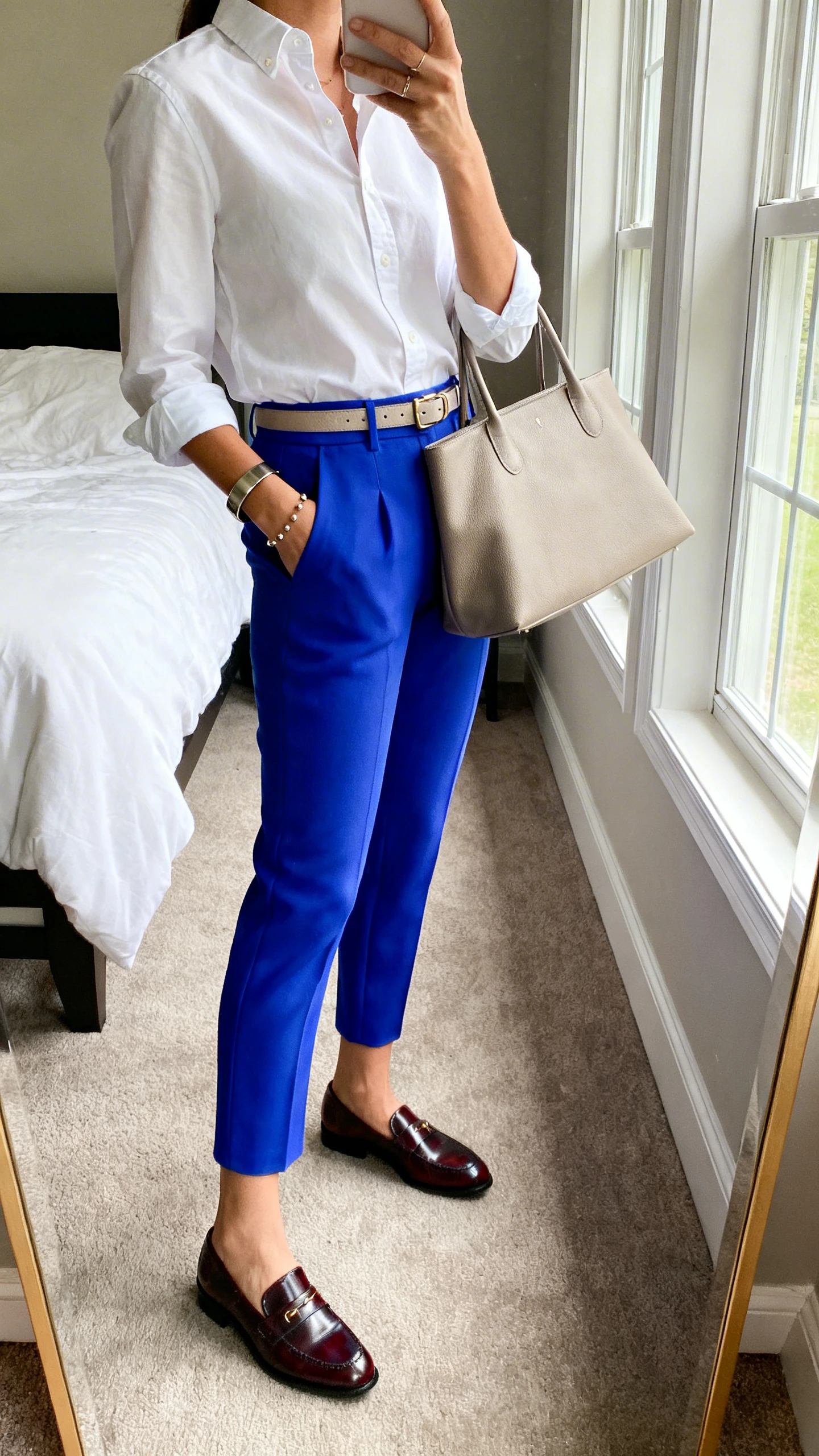 iPhone mirror selfie of a woman in vibrant cobalt ankle trousers with a crisp white button-down, polished loafers, a slim belt matching the shoes, understated studs, a sleek cuff bracelet, and a neutral structured tote, face not visible, side angle in a bedroom mirror, morning window light, iPhone photo quality.
