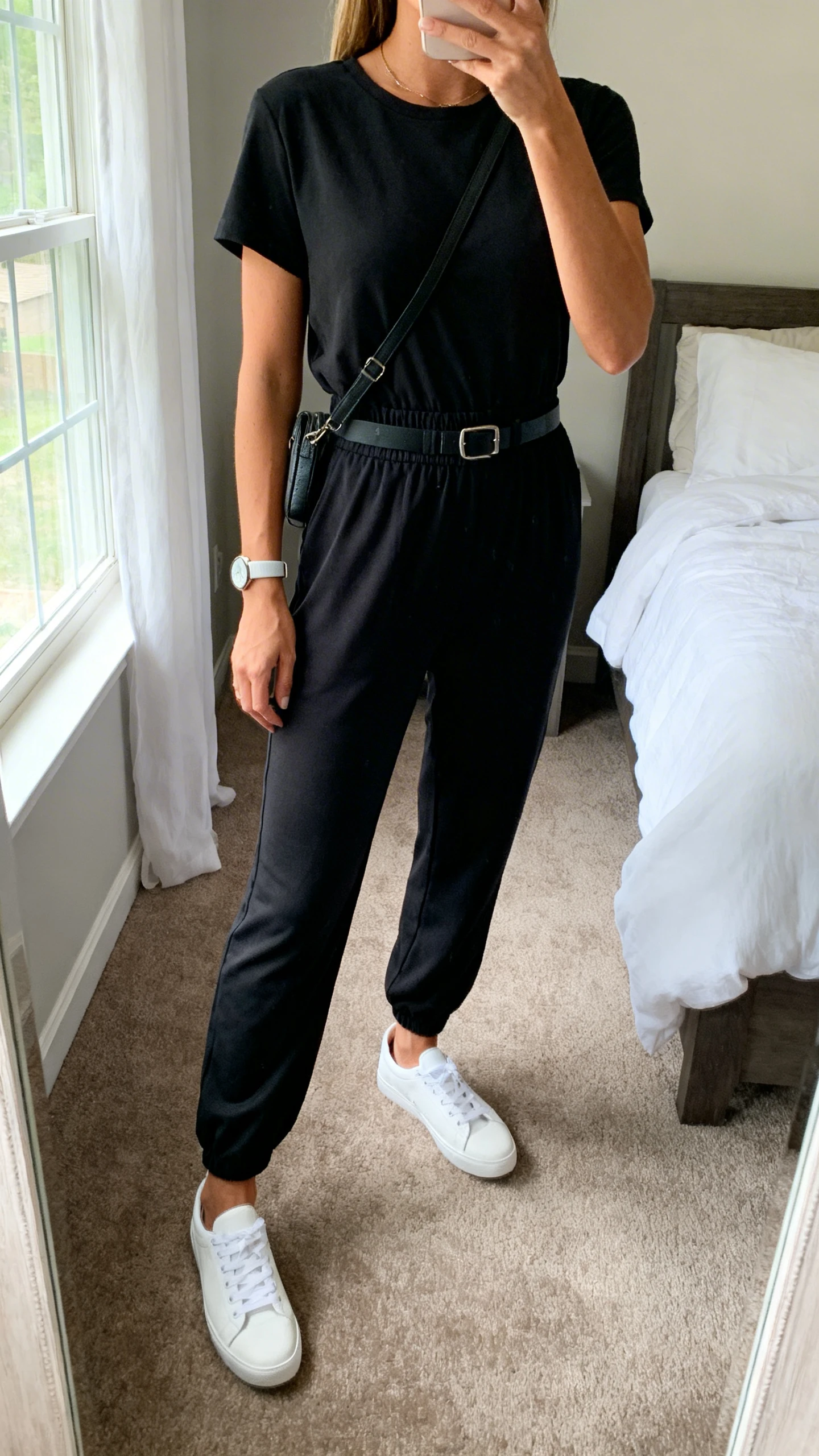iPhone mirror selfie of a woman wearing a black playproof stretch jumpsuit with short sleeves and elastic waist, white low-top sneakers, simple watch, and a crossbody belt bag, face not visible, cropped at neck, bedroom mirror, morning window light, iPhone photo quality.