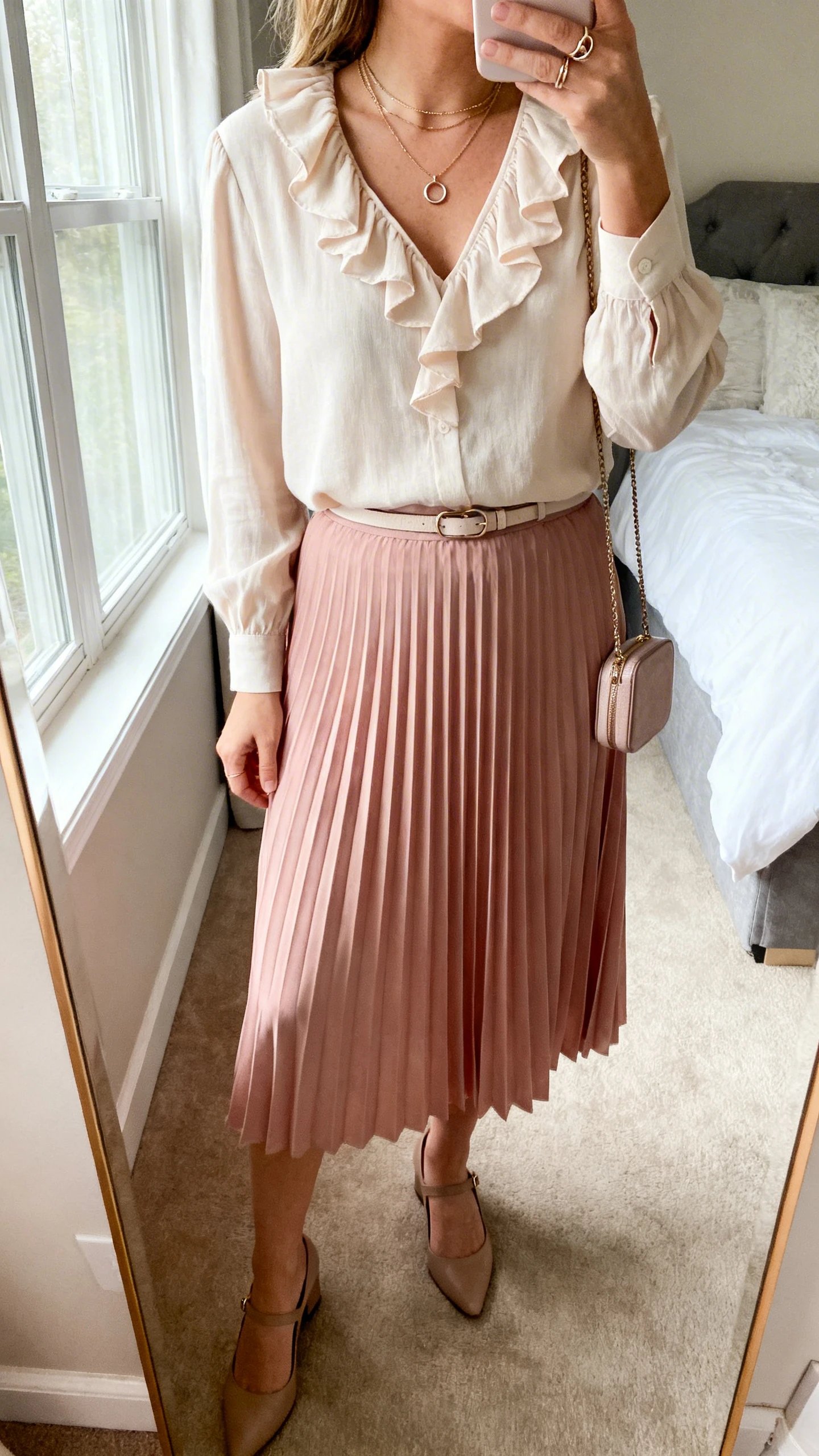 iPhone mirror selfie of a woman wearing a blush midi pleated skirt, a light ruffle-neck blouse tucked in, low slingback heels, a fine belt, layered delicate necklaces with a dainty ring stack, and a compact crossbody, face not visible, cropped at neck, bedroom mirror, soft window light, iPhone photo quality.