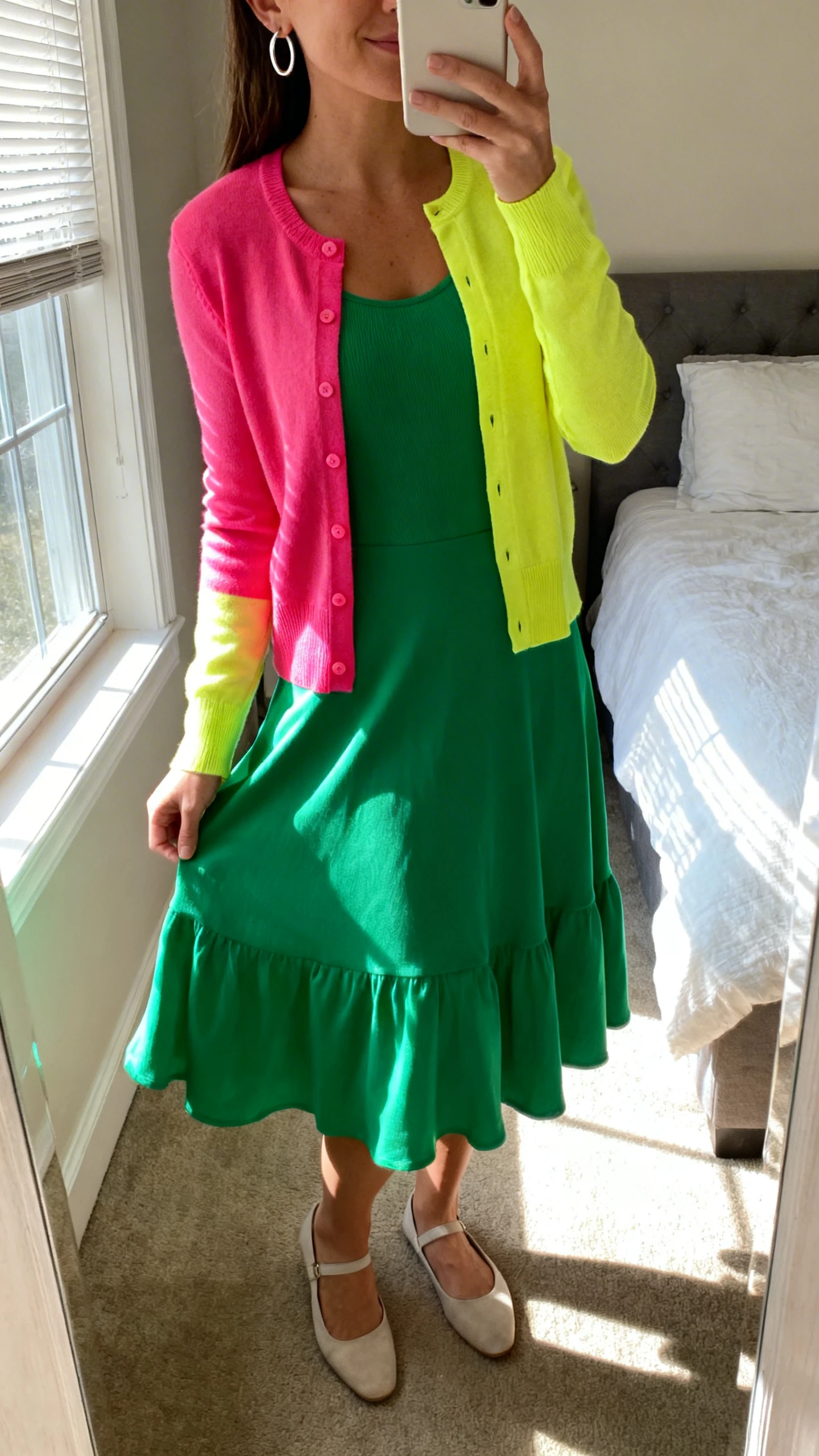 iPhone mirror selfie of a woman wearing a bright color-pop cardigan layered over a stretchy twirl-ready dress with simple hoop earrings and comfy low-profile flats, face not visible, cropped at neck, bedroom mirror, morning window light, iPhone photo quality.