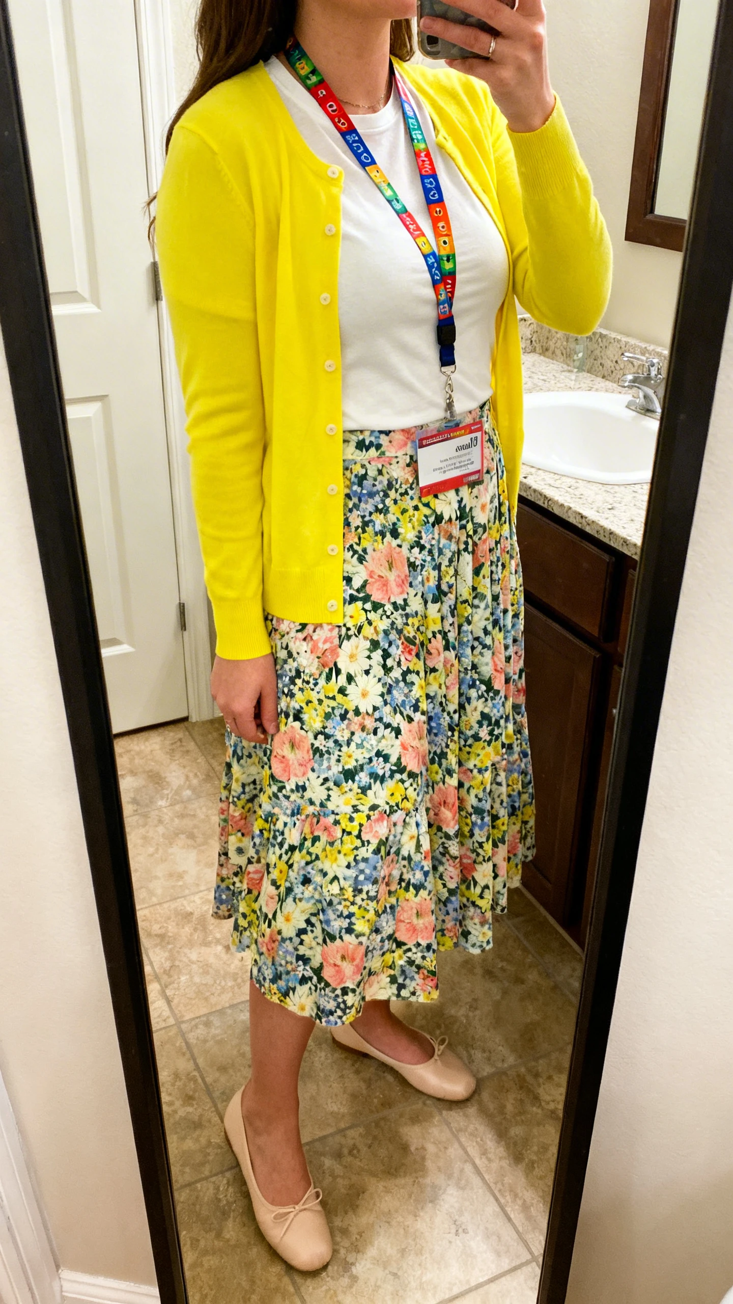 iPhone mirror selfie of a woman wearing a bright cardigan (sunny yellow) over a patterned midi skirt (whimsical floral print) with a white fitted tee, comfy ballet flats, and colorful teacher lanyard, face not visible, side angle, bathroom full-length mirror, soft indoor lighting, iPhone photo quality.
