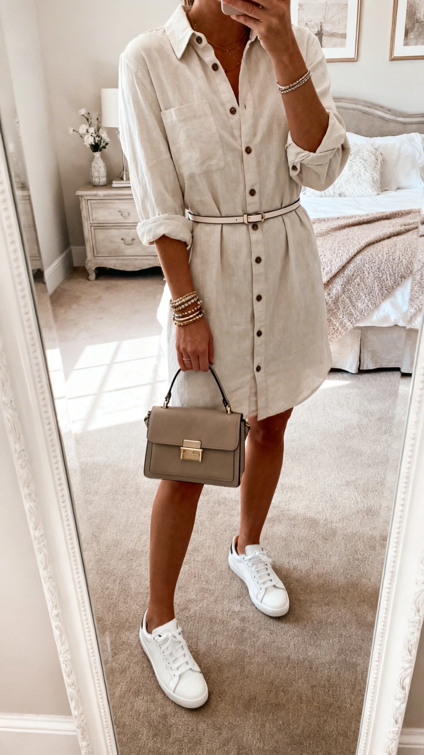 iPhone mirror selfie of a woman wearing a button-up shirt dress with rolled sleeves and a thin waist belt, white sneakers, layered bracelets, structured satchel, face not visible, cropped at shoulders, bedroom mirror, soft morning light, iPhone photo quality.