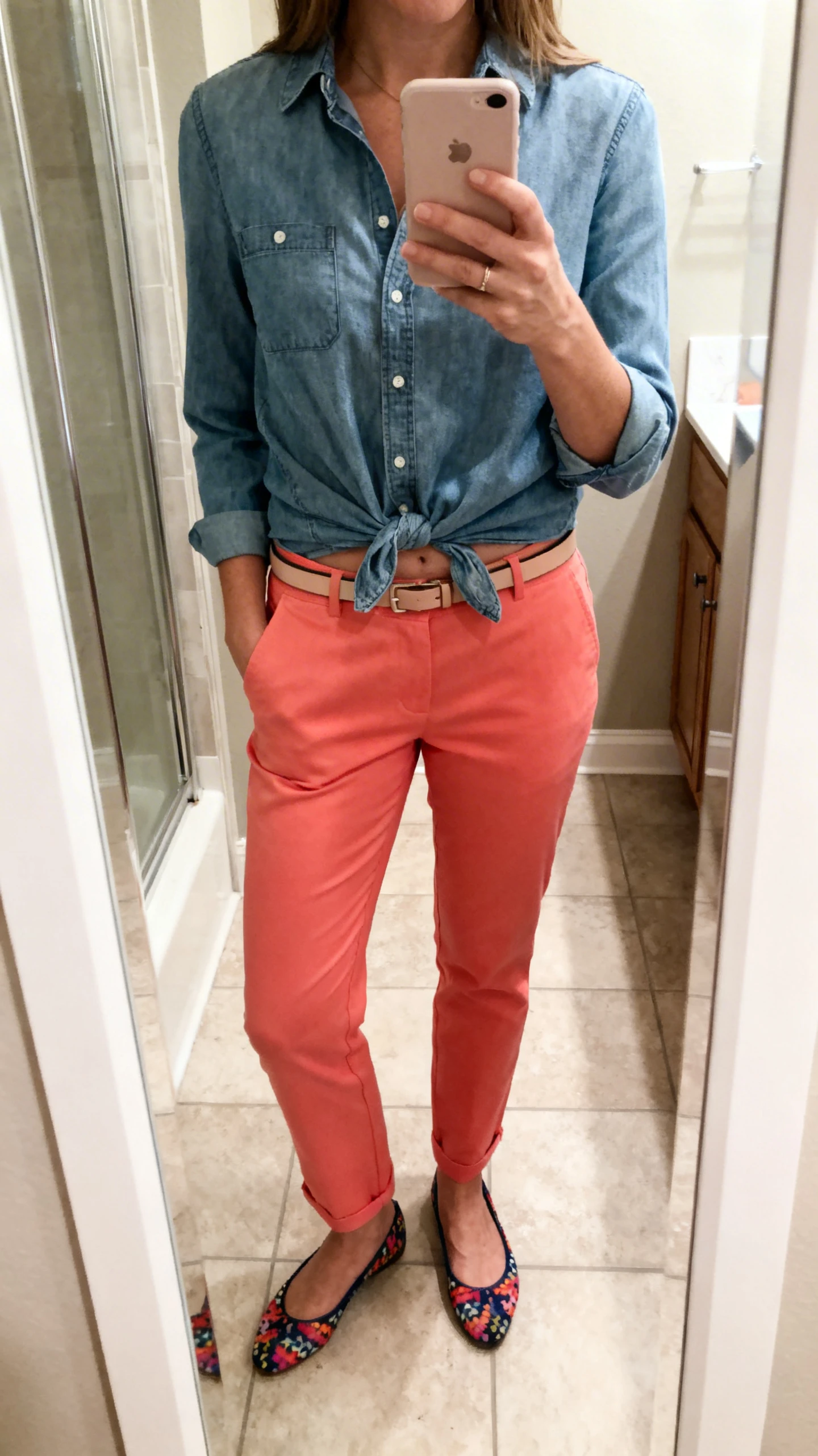 iPhone mirror selfie of a woman wearing a chambray shirt half-tucked into colored chinos with fun flats and a slim belt, face not visible, cropped at shoulders, bathroom mirror, soft indoor lighting, iPhone photo quality.