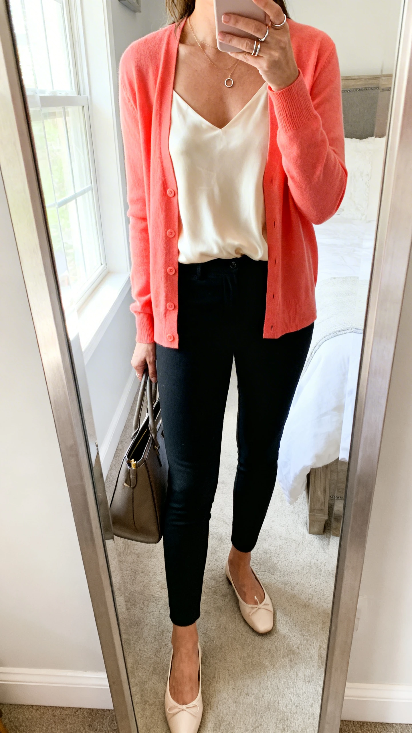 iPhone mirror selfie of a woman wearing a coral cardigan, black slim ankle pants, a cream silky shell top, classic ballet flats, delicate stacking rings with small hoops, and a structured work tote, face not visible, cropped at shoulders, full-length mirror in bedroom, bright window light, iPhone photo quality.