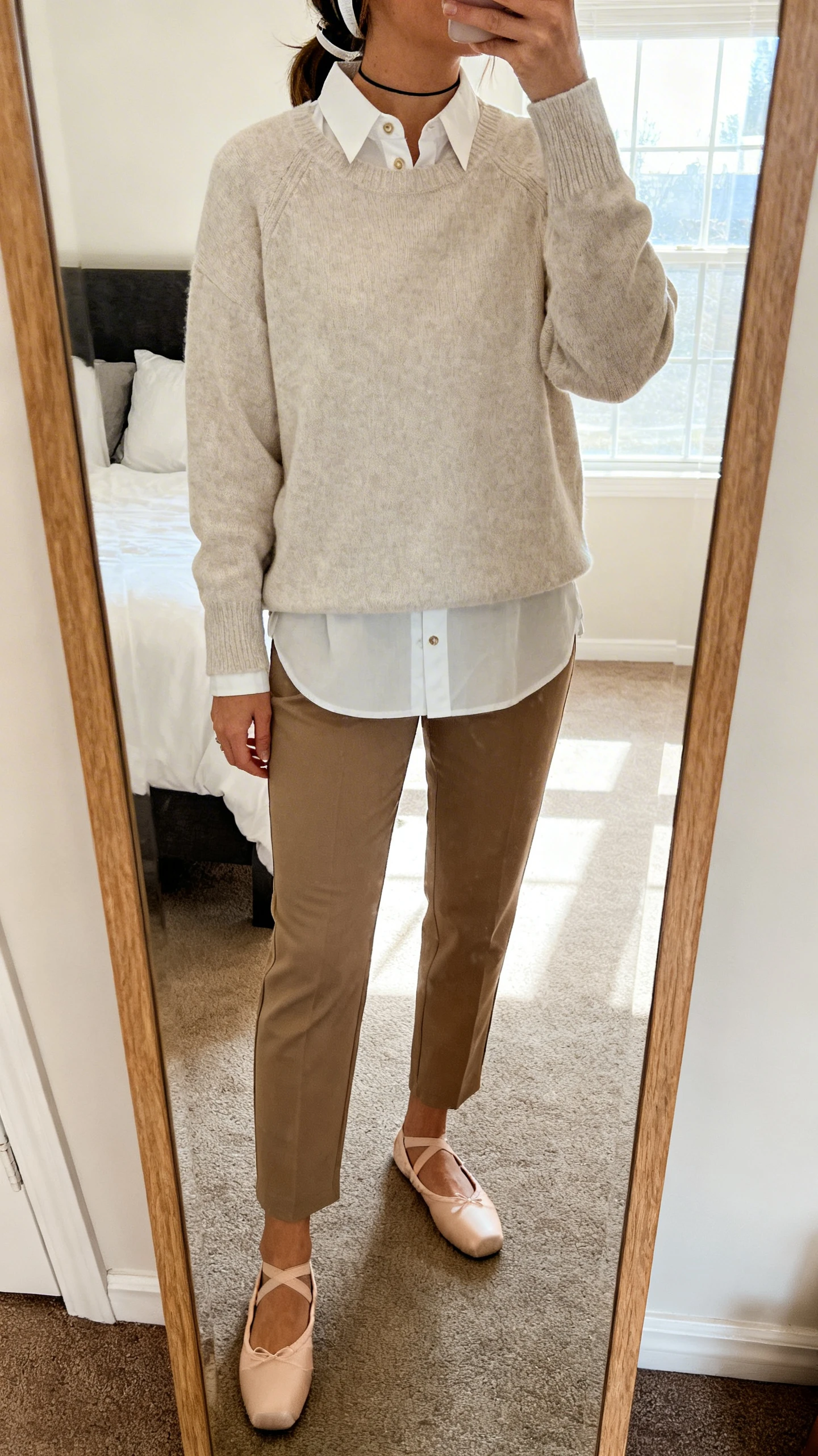 iPhone mirror selfie of a woman wearing a crisp button-down shirt layered under a lightweight crewneck sweater, straight-leg chinos, ballet flats, and a thin headband, face not visible, cropped at shoulders, bedroom mirror, natural window light, iPhone photo quality.
