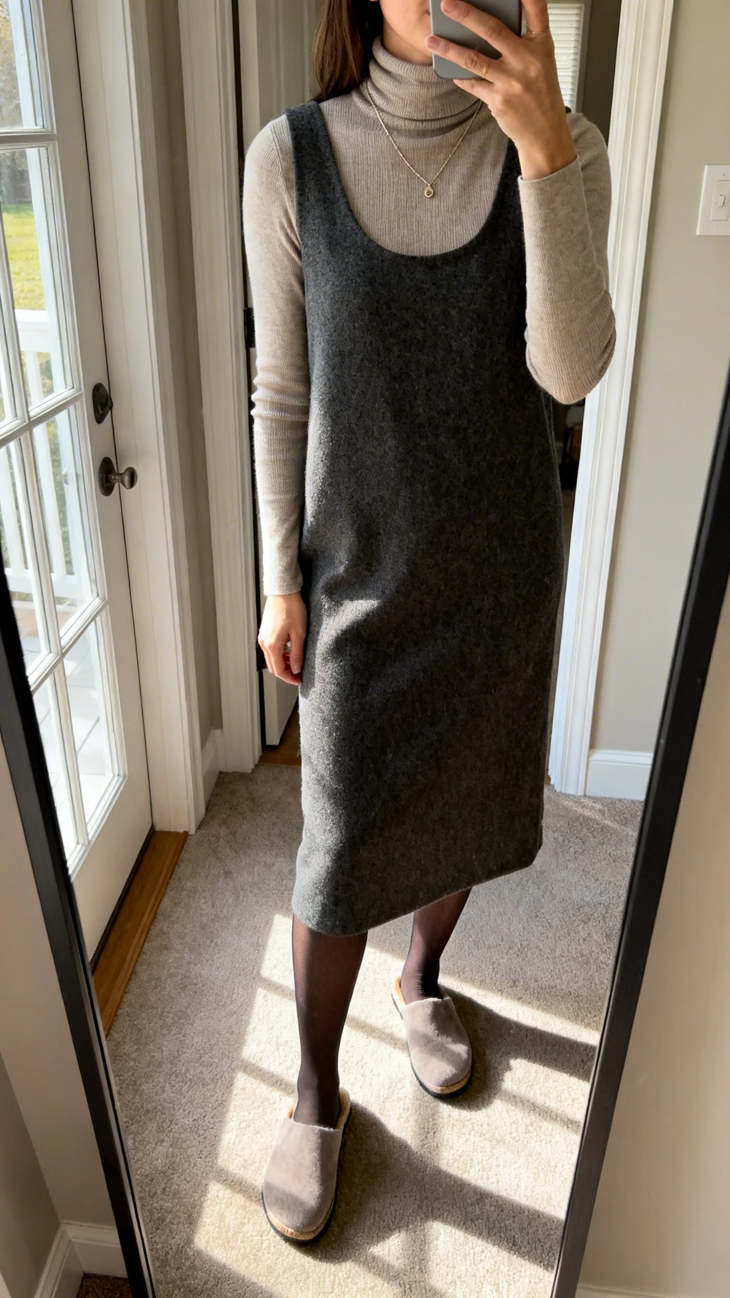 iPhone mirror selfie of a woman wearing a fitted turtleneck under a sleeveless midi dress with opaque tights and comfy mules, delicate necklace, face not visible, cropped at shoulders, entryway mirror, morning window light, iPhone photo quality.