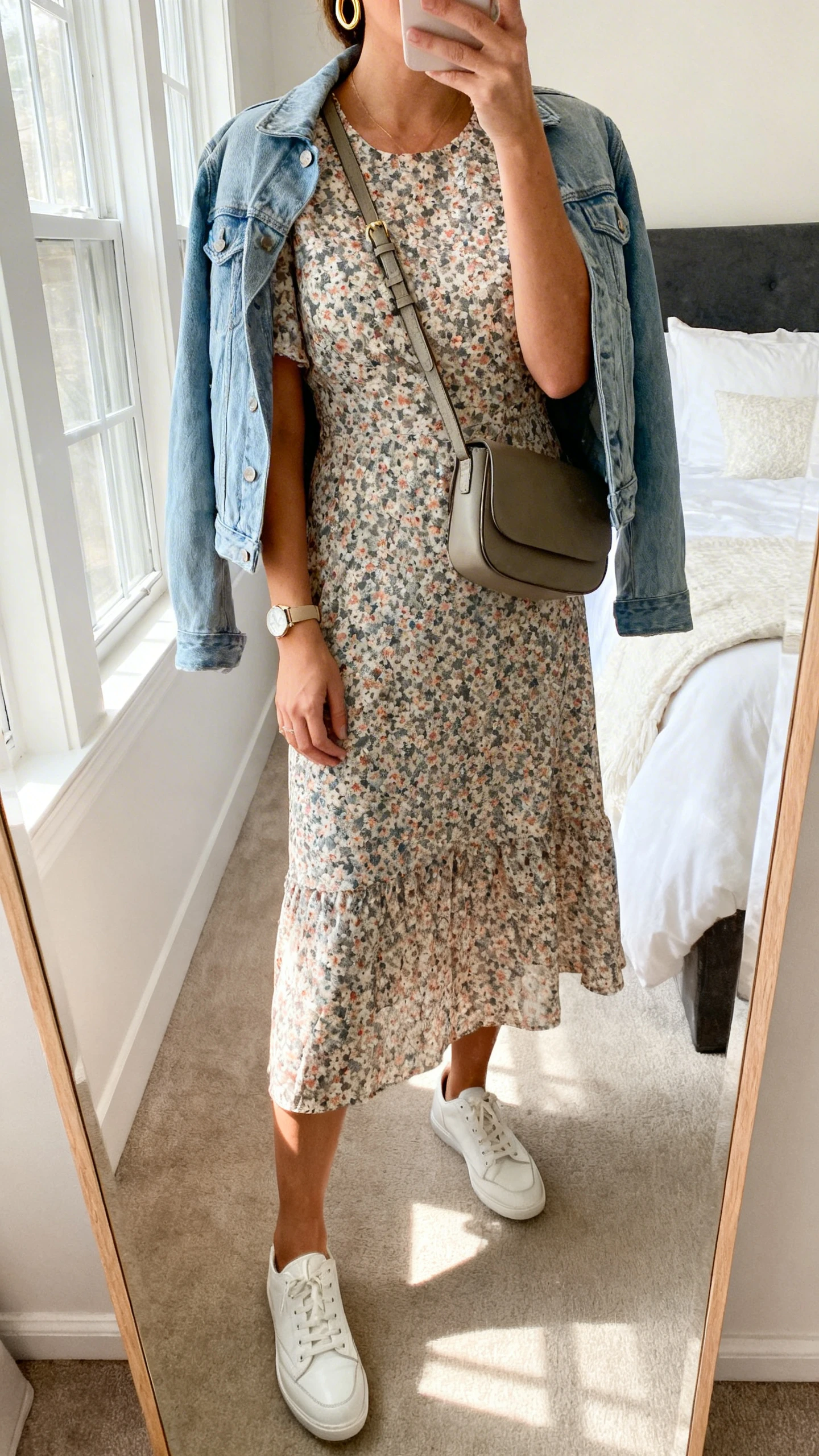 iPhone mirror selfie of a woman wearing a floral midi dress with short sleeves, clean white leather sneakers, a lightweight denim jacket, simple gold hoops, a structured crossbody bag, and a subtle watch, face not visible, cropped at neck, bedroom mirror, soft morning window light, iPhone photo quality.