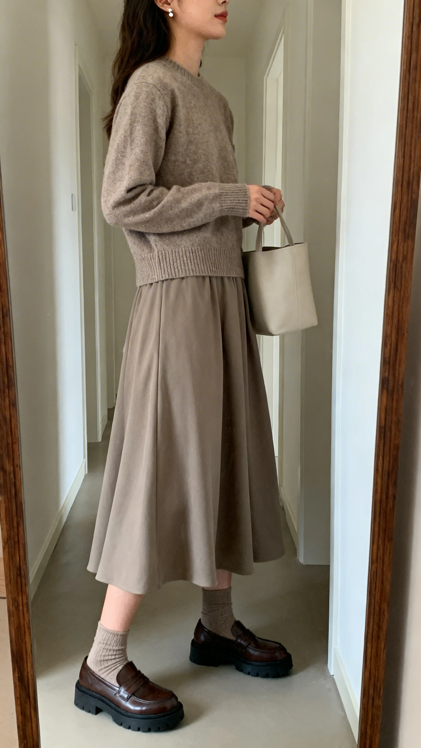 iPhone mirror selfie of a woman wearing a flowy midi skirt, tucked-in crewneck sweater, chunky loafers with socks, simple tote and delicate studs, face not visible, side angle in a hallway full-length mirror, soft indoor lighting, iPhone photo quality
