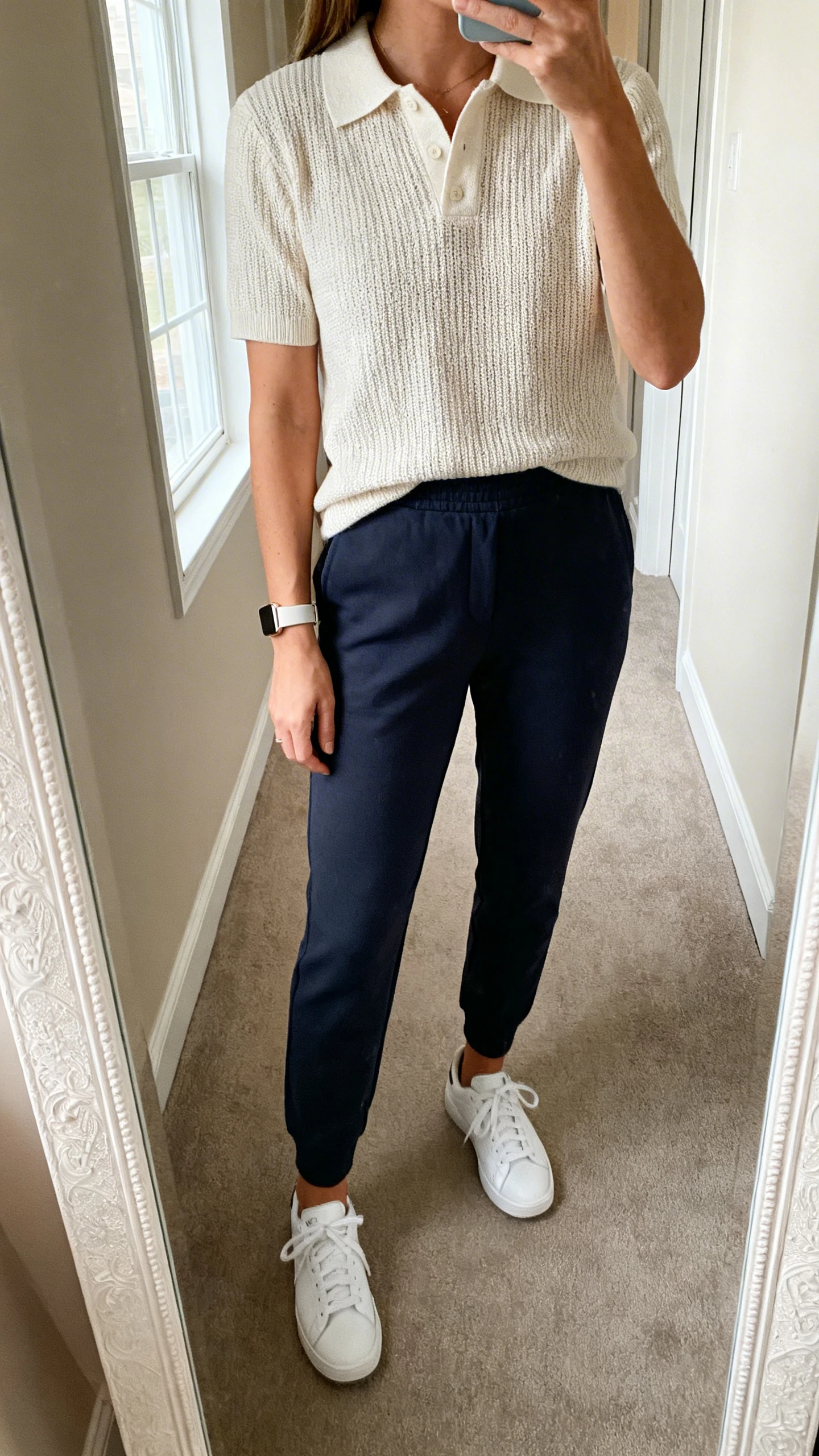 iPhone mirror selfie of a woman wearing a knit polo tucked into pull-on pants with clean white sneakers and a minimalist watch, face not visible, cropped at shoulders, hallway mirror, soft window light, iPhone photo quality.