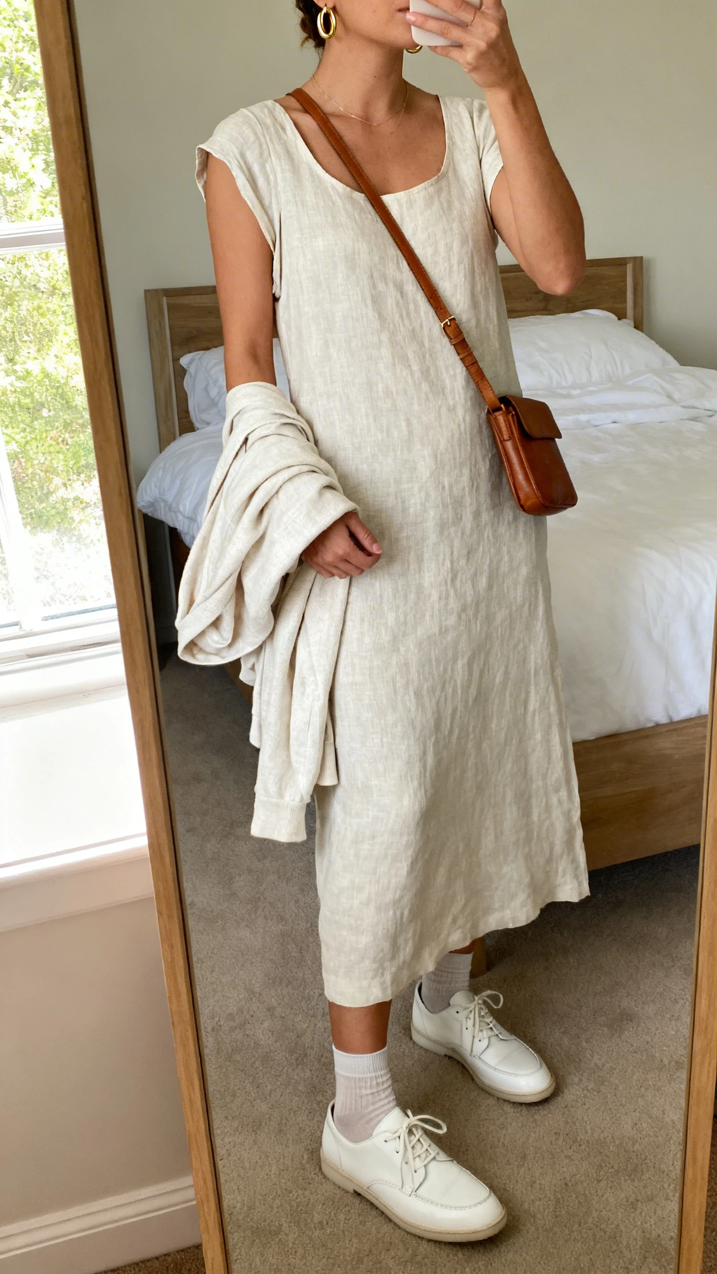 iPhone mirror selfie of a woman wearing a lightweight linen midi dress (tank/short sleeves) with clean white leather sneakers, delicate gold hoop earrings, a simple leather crossbody, breathable no-show socks, and a light cotton cardigan draped over her arm, face not visible, cropped at neck, bedroom mirror, soft morning window light, iPhone photo quality.
