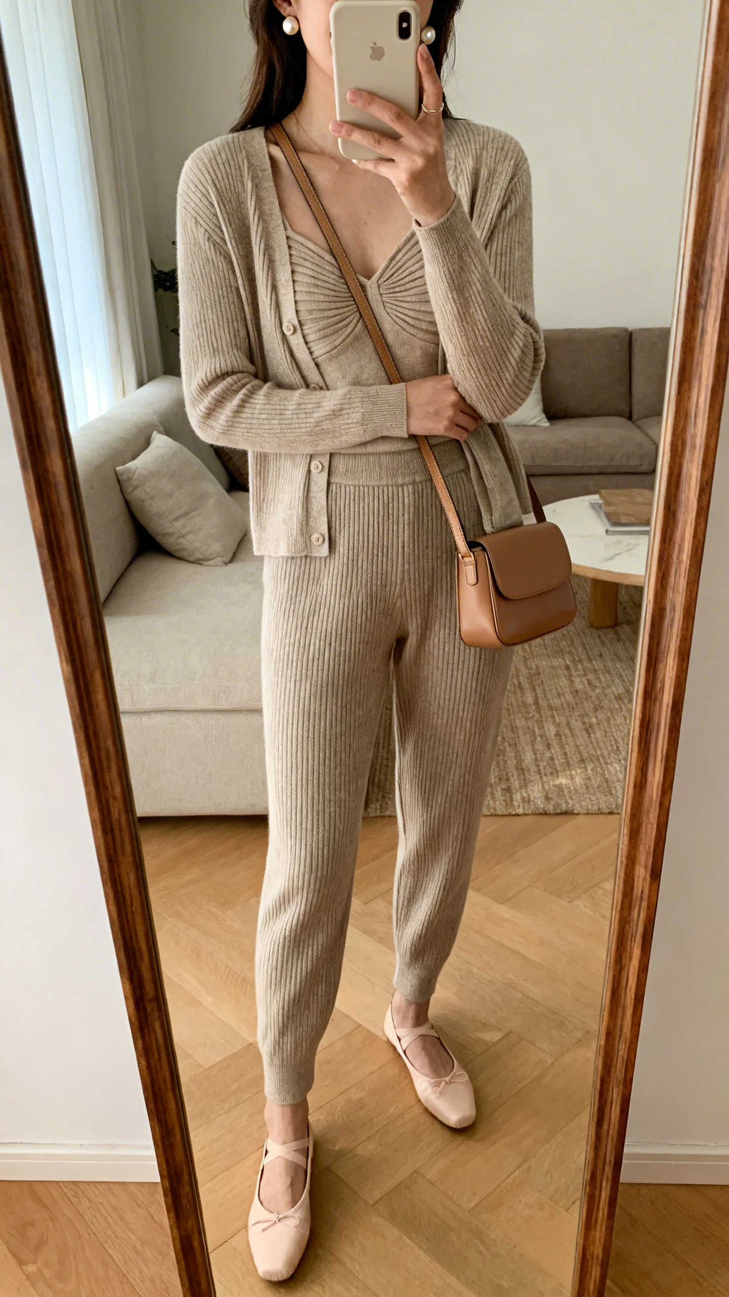 iPhone mirror selfie of a woman wearing a matching cardigan set (shell top and cardigan) tucked into high-waisted knit pants, ballet flats, pearl studs, slim leather shoulder bag, face not visible, cropped at neck, living room full-length mirror, warm indoor lighting, iPhone photo quality.