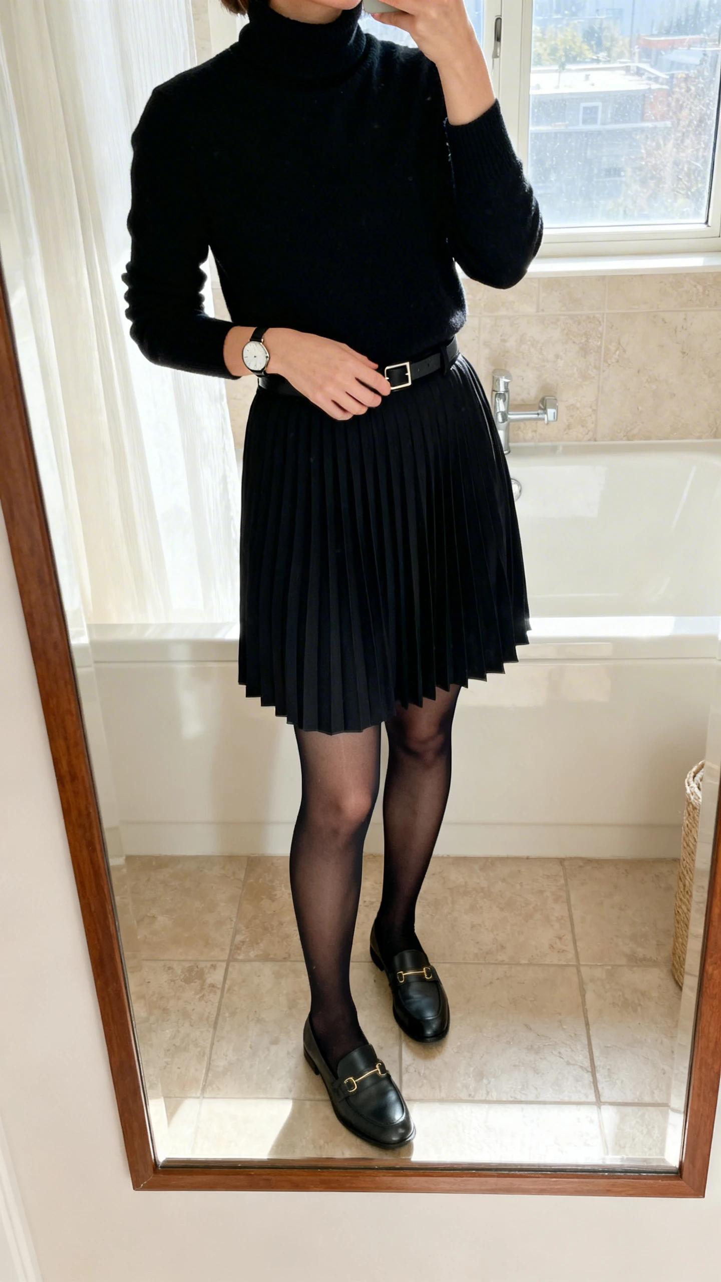 iPhone mirror selfie of a woman wearing a monochrome black turtleneck tucked into a black pleated skirt, sheer black tights, loafers, minimalist watch, face not visible, cropped at shoulders, bathroom mirror, natural window light, iPhone photo quality