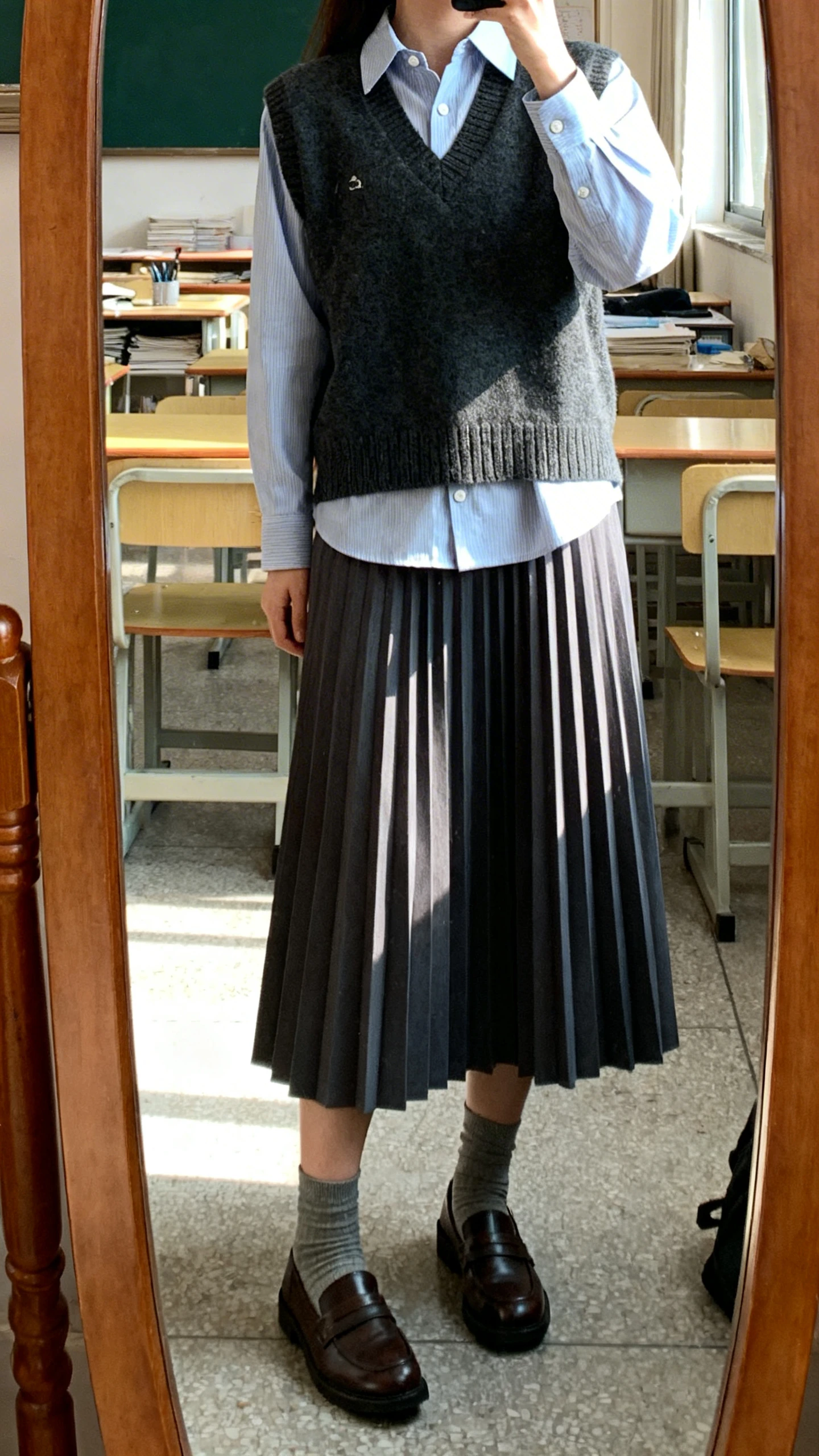 iPhone mirror selfie of a woman wearing a pleated midi skirt with a button-up shirt under a sweater vest, paired with loafers and ankle socks, face not visible, cropped at neck, classroom-style home office mirror, indoor light, iPhone photo quality.