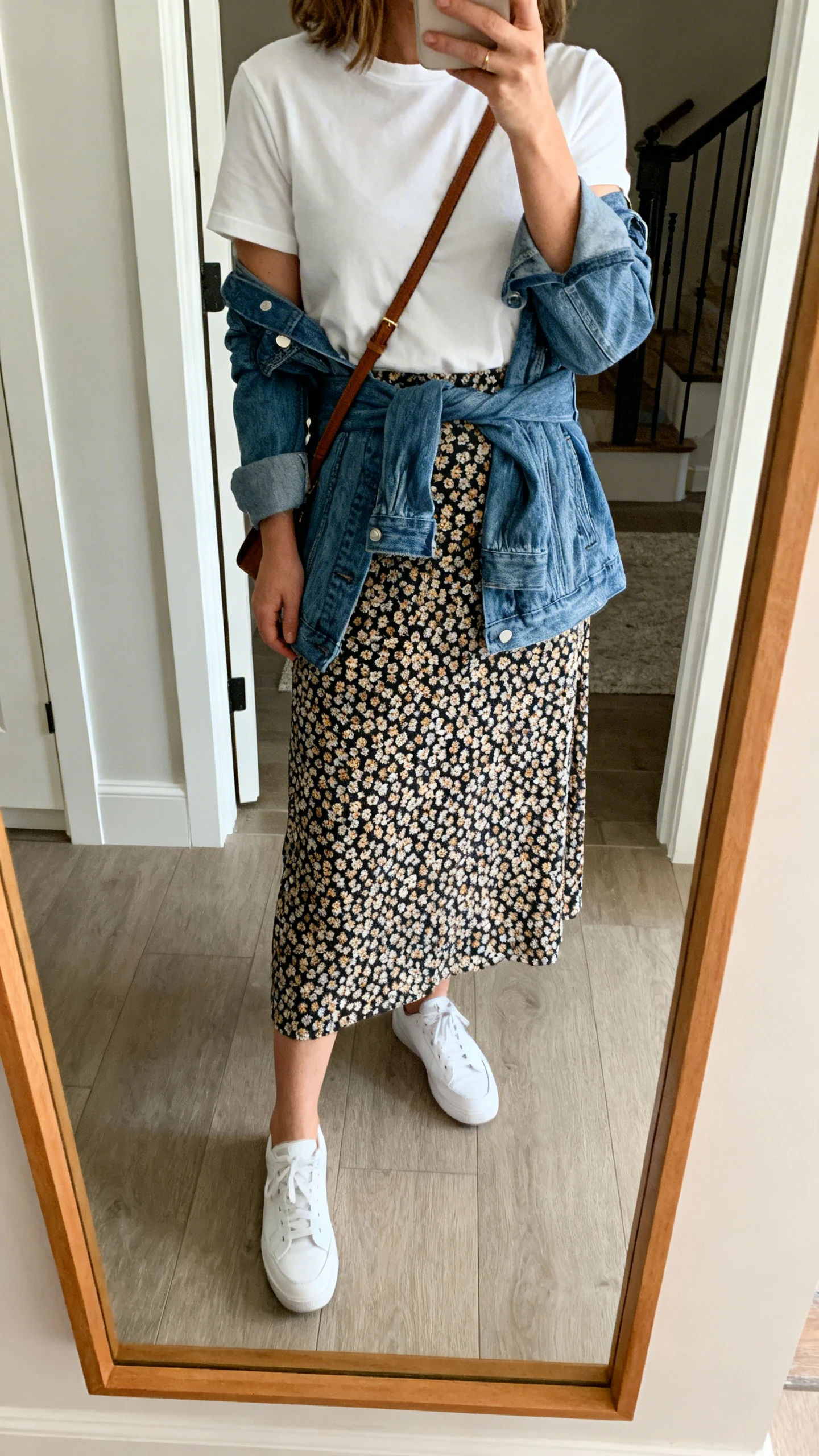 iPhone mirror selfie of a woman wearing a printed midi skirt with white sneakers, tucked-in crewneck tee, denim jacket tied at the waist, crossbody bag, face not visible, cropped at shoulders, entryway mirror, natural daylight, iPhone photo quality.