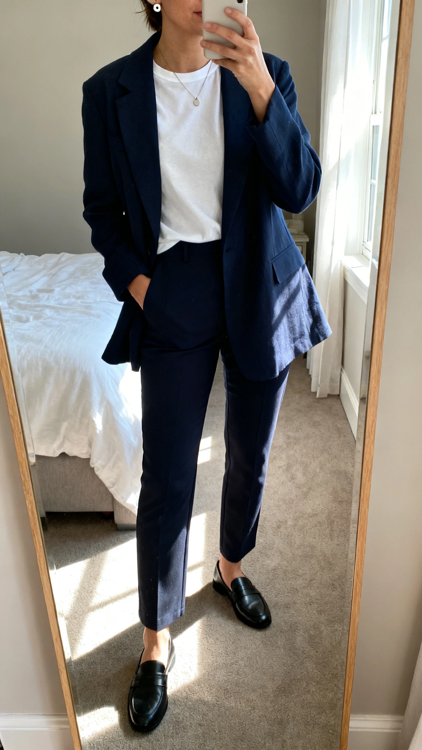 iPhone mirror selfie of a woman wearing a relaxed-fit navy blazer over a white cotton crewneck tee, tailored ankle-length trousers with stretch, black loafers, a minimal pendant necklace and stud earrings, face not visible, cropped at neck, bedroom mirror with morning window light, iPhone photo quality.