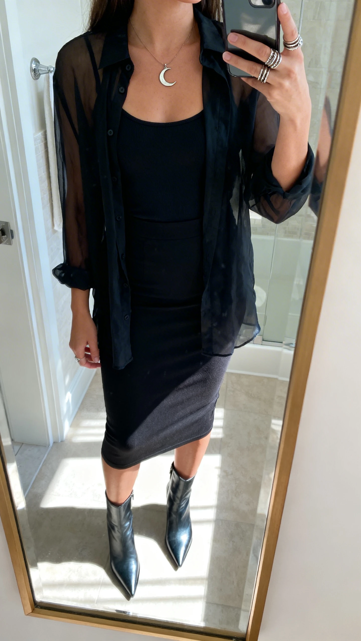 iPhone mirror selfie of a woman wearing a sheer black blouse over a black camisole, fitted black pencil skirt, pointed ankle boots, witchy details like a crescent moon pendant and stackable rings, face not visible, cropped at neck, bathroom mirror, natural morning light, iPhone photo quality