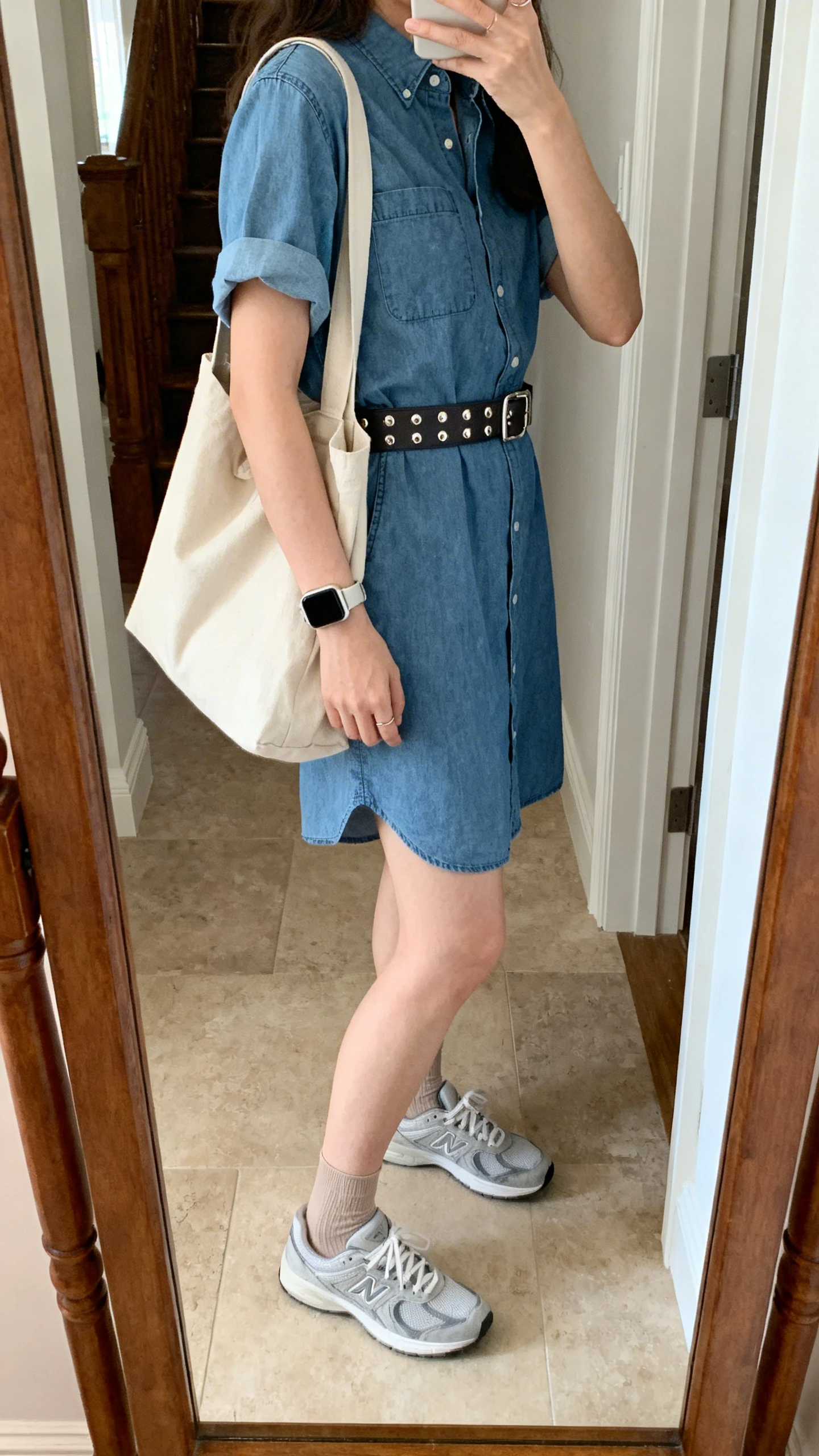 iPhone mirror selfie of a woman wearing a short-sleeve shirt dress (cotton/chambray) with clean sneakers, a wide waist belt (optional), polished studs, a smartwatch, a large canvas tote, and thin ankle socks matching shoes, face not visible, side angle in an entryway mirror with natural daylight, iPhone photo quality.