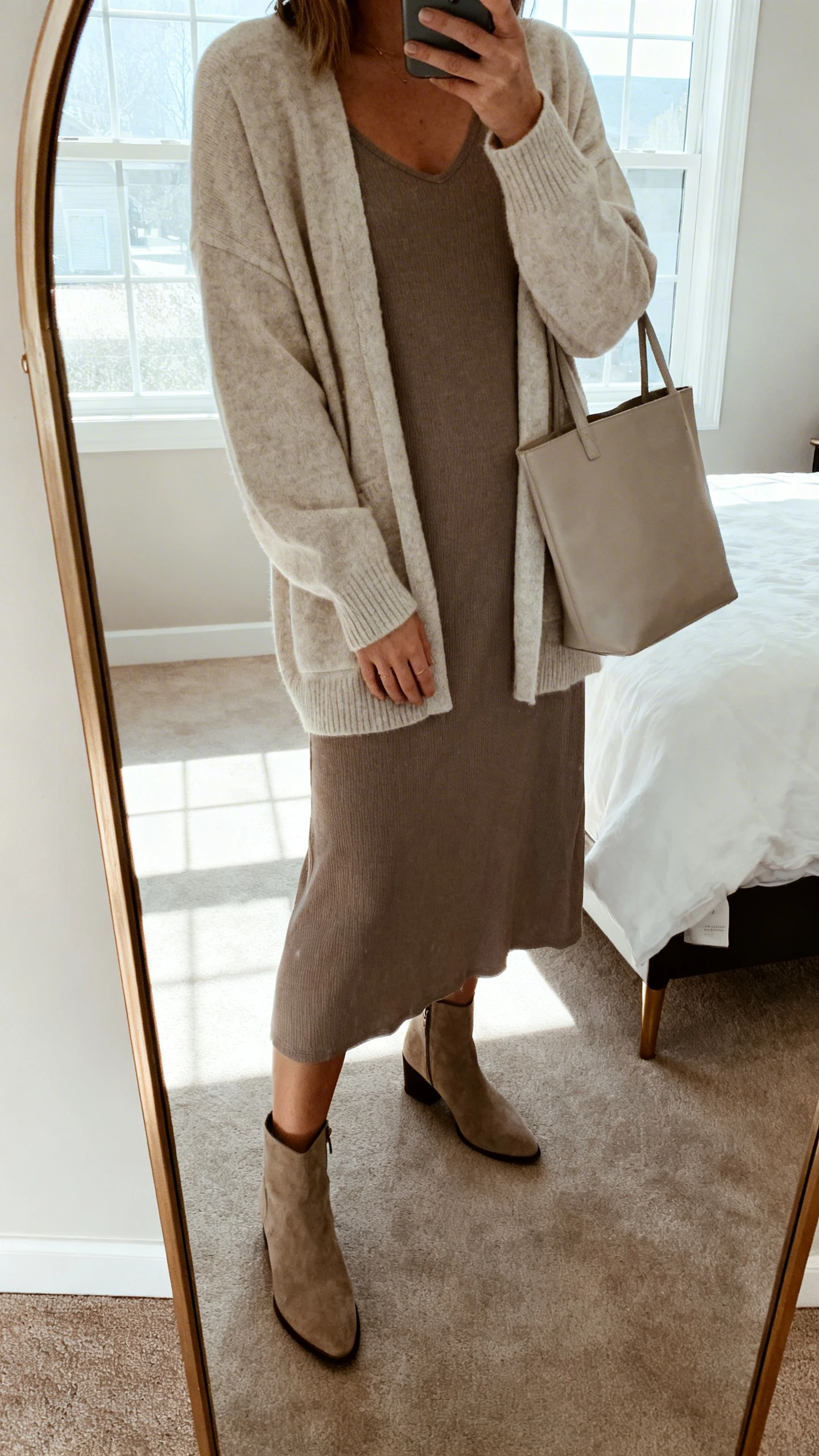 iPhone mirror selfie of a woman wearing a soft cardigan layered over a midi dress with ankle boots and a simple tote, face not visible, cropped at neck, bedroom mirror, natural window light, iPhone photo quality