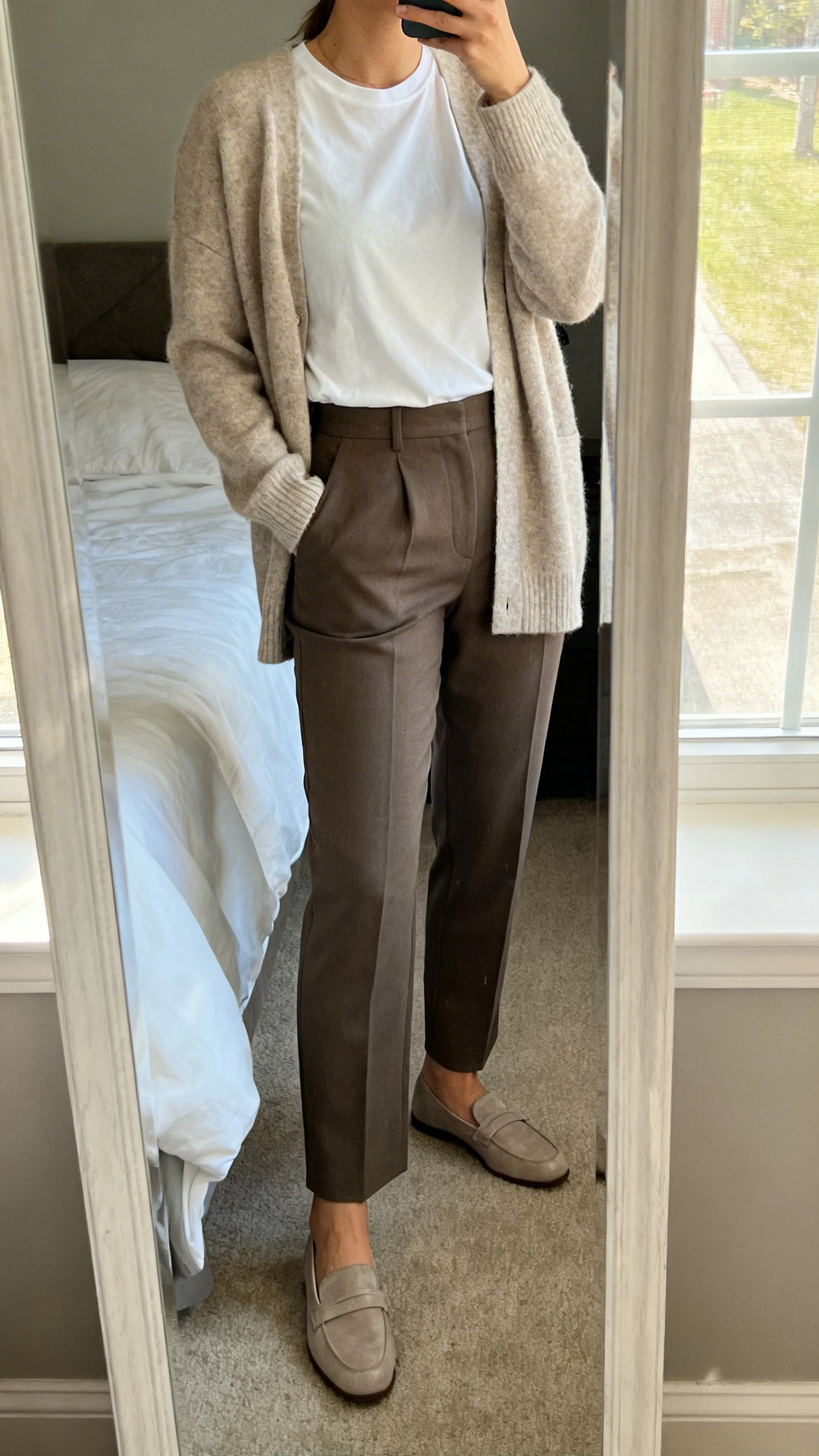 iPhone mirror selfie of a woman wearing a soft cardigan layered over a classic tee with tailored trousers and simple loafers, face not visible, cropped at neck, bedroom mirror, natural window light, iPhone photo quality.