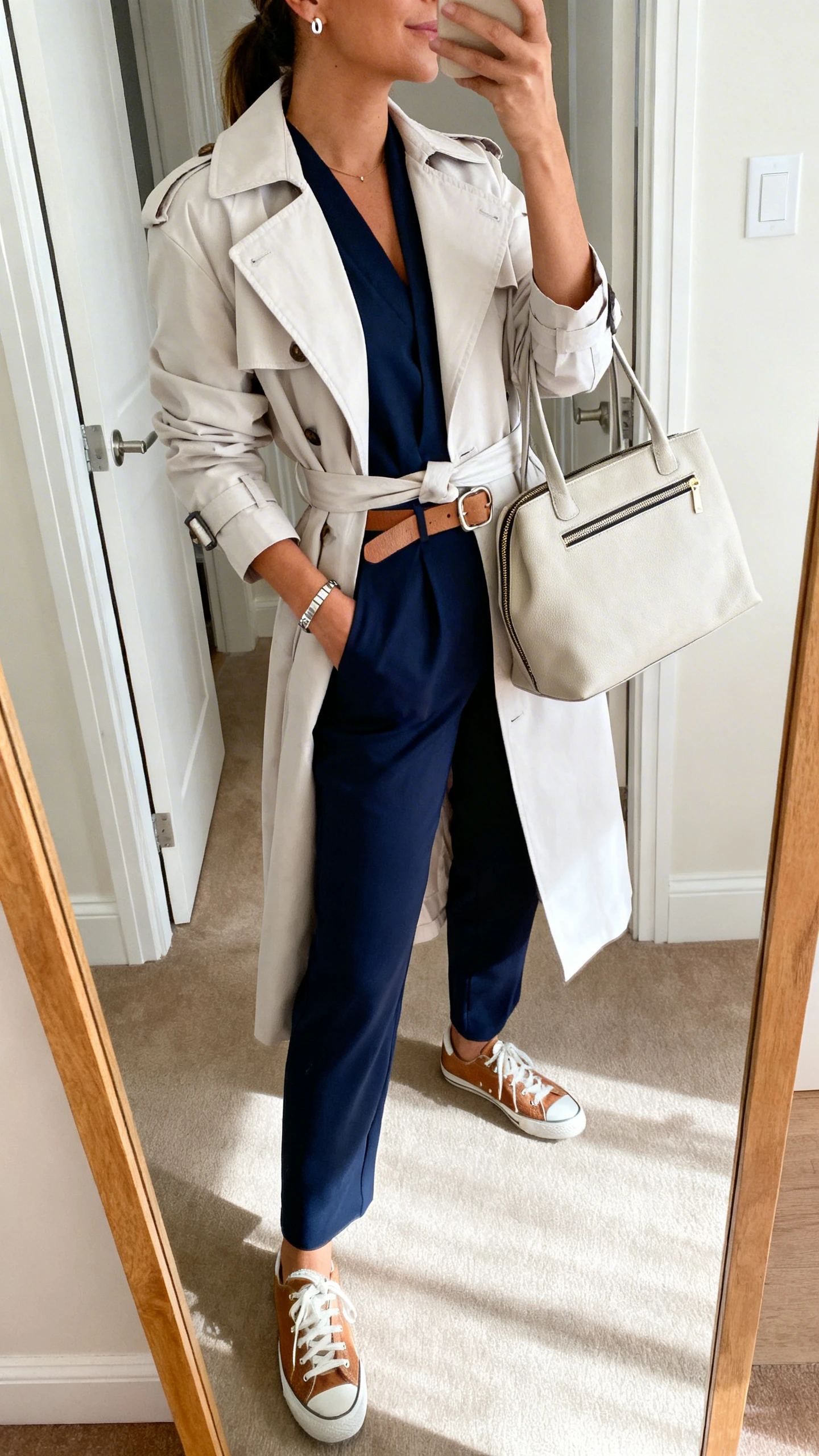 iPhone mirror selfie of a woman wearing a tailored navy jumpsuit under a light belted trench, leather sneakers, simple belt, stud earrings with a sleek bracelet, and a zip-top tote, face not visible, side angle, entryway mirror, bright morning light, iPhone photo quality.